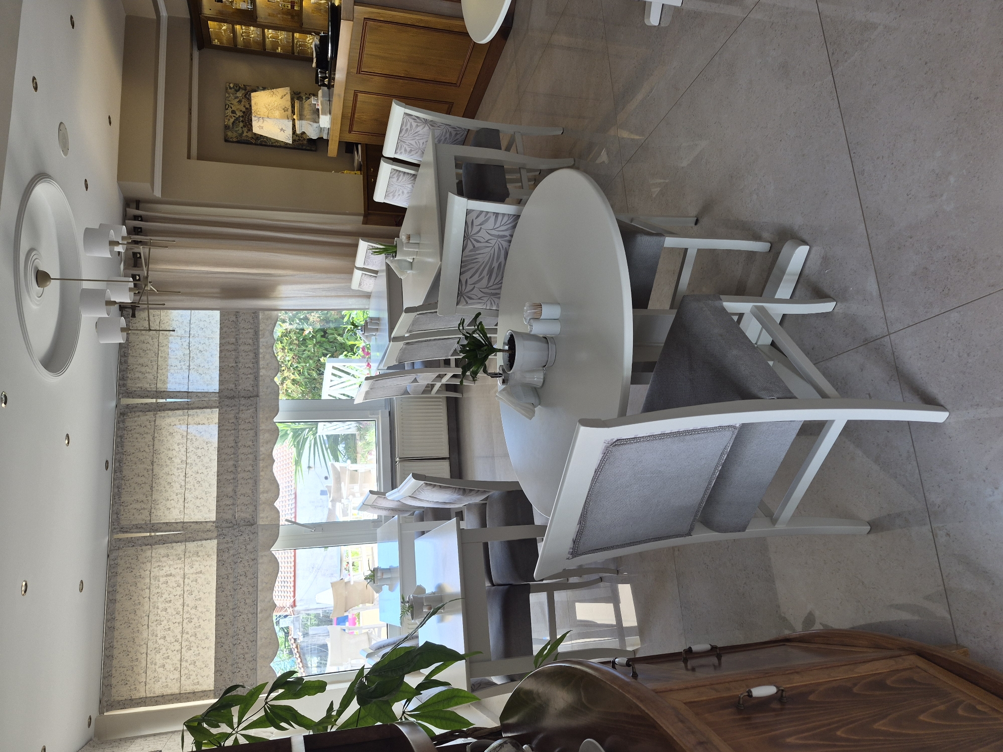 The image depicts the interior of a restaurant or cafe, likely located in Νέα Καλλικράτεια, Greece, based on the context provided. The scene is brightly lit, with a spacious dining area furnished with several white tables and chairs. The tables are round and rectangular, some set with salt and pepper shakers and toothpicks, indicating readiness for diners. The chairs are upholstered in grey fabric with decorative patterns on the backrests.
Large windows with patterned Roman blinds line one wall, offering a view of lush greenery and outdoor seating areas, suggesting a pleasant, possibly seaside, location. The floor is tiled with a polished, light-colored material that reflects the overhead lighting.
In the background, a wooden bar area is visible, featuring a display of glassware and a prominent lamp with a patterned shade. A large potted plant is positioned in the foreground, adding a touch of nature to the indoor environment. The overall ambiance is clean, modern, and inviting. There are no people visible in the image.