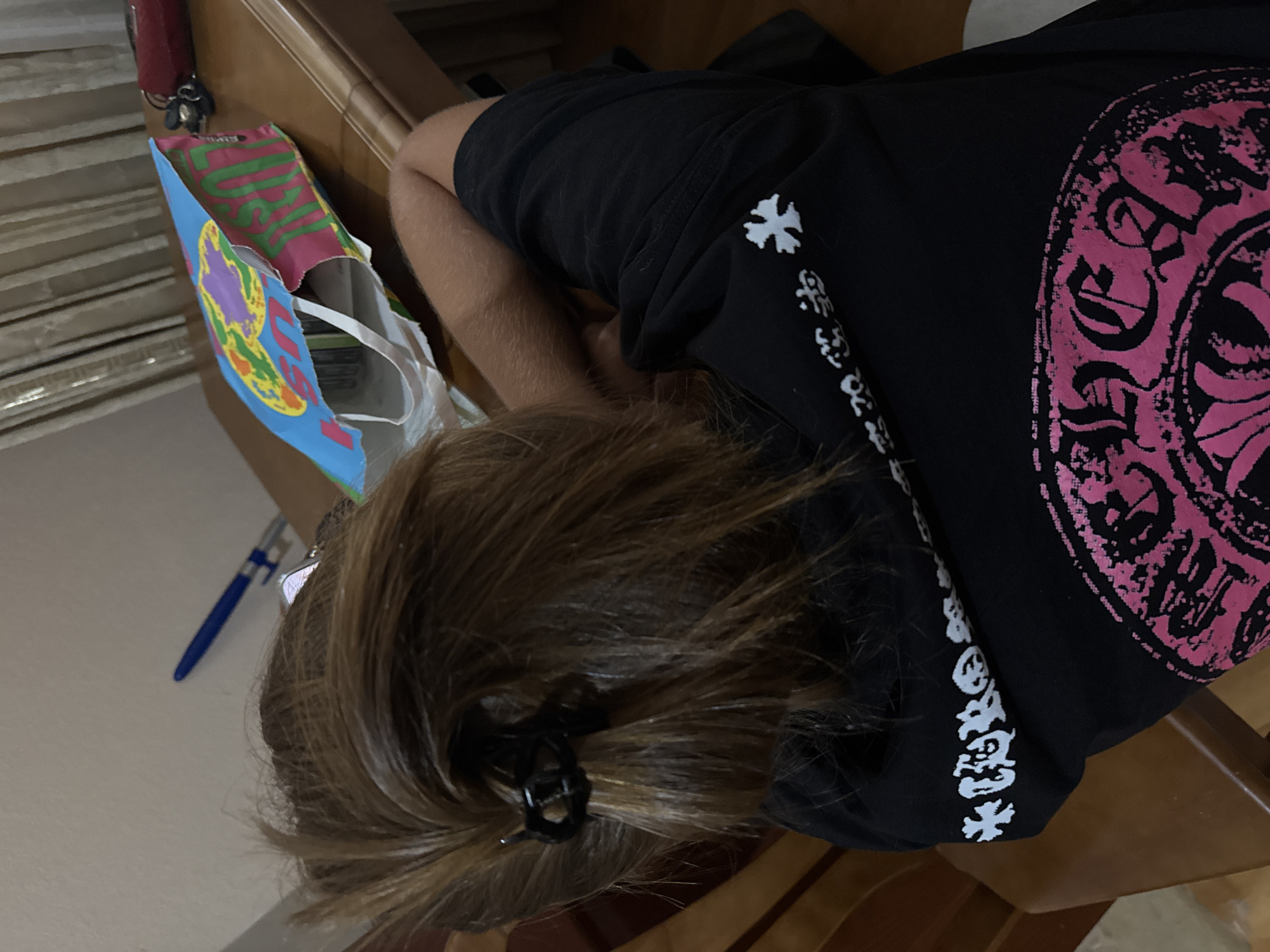 A person with light brown hair, styled in a messy bun held by a black hair clip, is seated at a wooden table. They are wearing a black t-shirt with a white graphic print on the back. The graphic features a floral or ornamental design in pink and white. To the left of the person, a blue pen is visible on the table. In front of the person is an open paper bag with a colorful, abstract design, appearing to be a shopping bag. A portion of a red object, possibly a wallet or clutch, is also on the table. The background shows vertical blinds and a wall. The scene is indoors, likely in a residential setting.