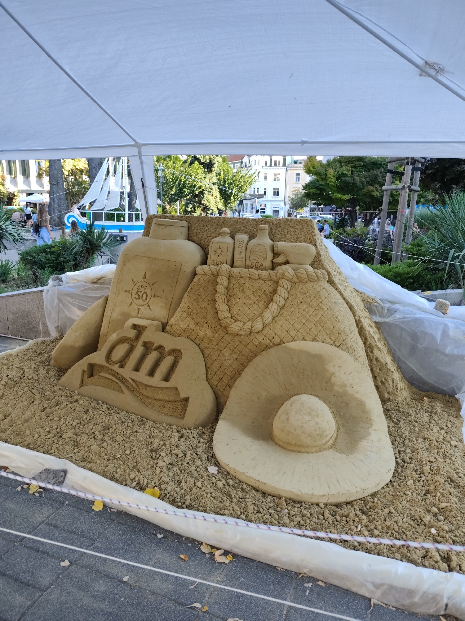 The image depicts a sand sculpture in the city of Burgas, Bulgaria, located at the Sea Garden. The sculpture is situated under a white tent, with a cobblestone walkway visible in the foreground. The sand sculpture prominently features a large bottle of sunscreen with "SPF 50" inscribed on it, a woven beach bag, and a wide-brimmed hat, evoking a summer beach theme. To the left of the sunscreen bottle, a sculpted logo for "dm" (a drugstore chain) is visible. In the background, trees, buildings, and people can be observed, along with a decorative boat structure. The overall scene suggests an outdoor public display or event.