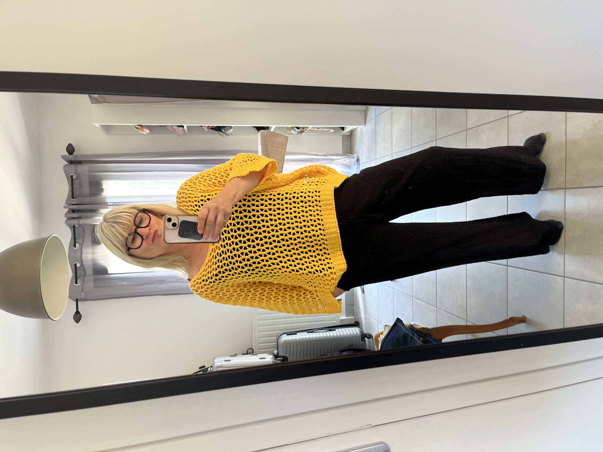 The image shows a woman taking a mirror selfie in what appears to be a residential setting. She is standing in front of a full-length mirror, wearing a yellow crocheted sweater and black wide-leg pants. She has blonde hair and is wearing glasses. The background includes a lamp, curtains, a closet, and luggage. The floor is tiled. There is no indication of the city of Chambœuf, France.