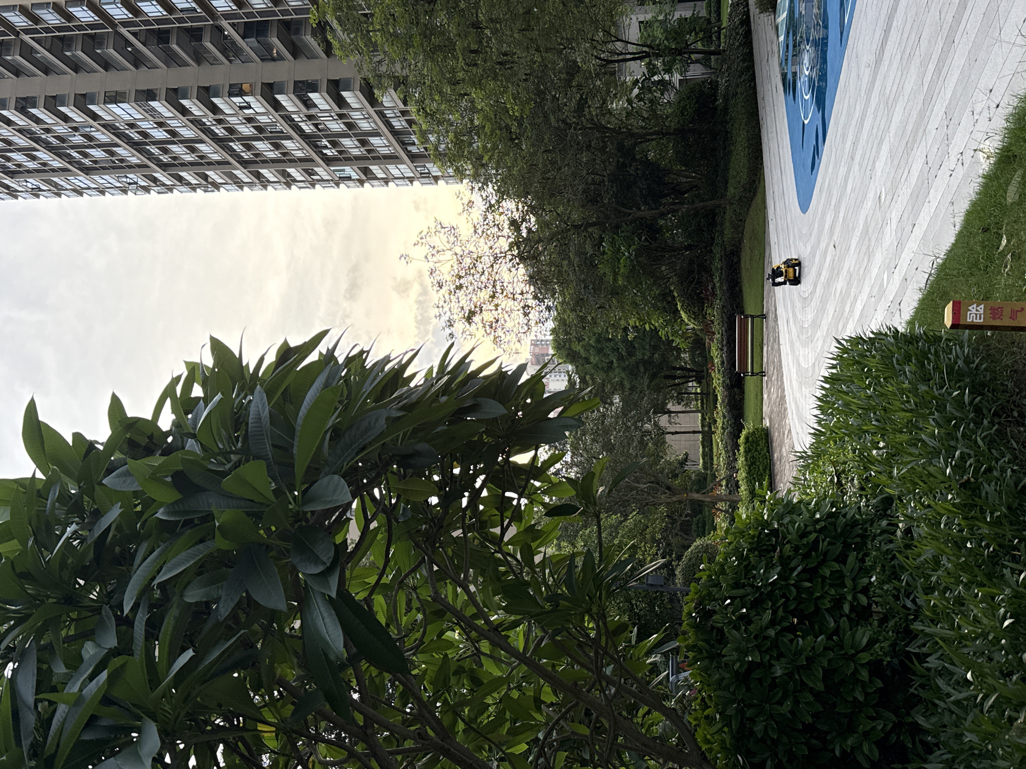 The image depicts a residential complex in Huizhou, China, featuring a modern high-rise building and a landscaped garden. A child is seen riding a yellow toy vehicle on a paved pathway. The pathway is bordered by lush green bushes and trees, and a park bench is visible in the background. A portion of a blue and white play area is also present. The sky is overcast with the sun partially visible, casting a warm light on the scene. The overall impression is one of a tranquil urban environment designed for recreation and family living.
