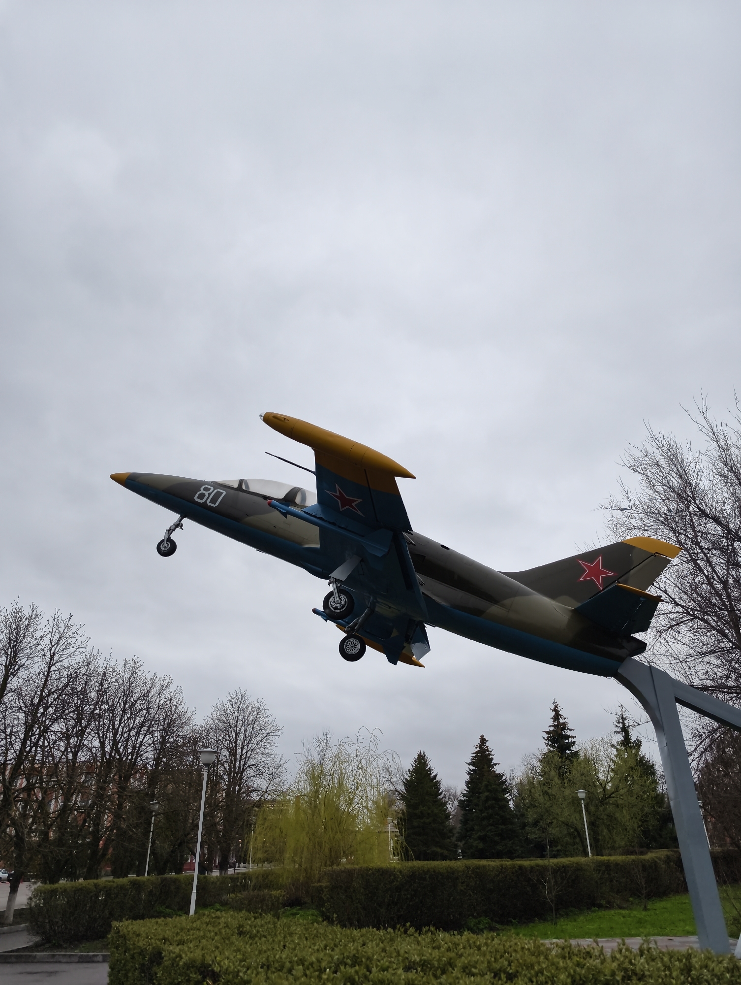 This media file displays a jet aircraft, likely a trainer or light attack model, mounted on a grey metal stand in a public park in Balashov, Russia. The aircraft is angled upwards, as if in flight, and its landing gear is extended. It features a camouflage paint scheme of dark grey/green on the upper fuselage and blue on the underside, accented with yellow tips on the wings and tail. The number "80" is clearly visible on the side of the fuselage near the cockpit, and a red star insignia, characteristic of Soviet/Russian military aircraft, is emblazoned on the vertical stabilizer.
The scene is set outdoors under an overcast sky, indicating a cloudy daytime. The park environment includes well-maintained green hedges and grass in the foreground. In the background, a mix of trees is present: bare deciduous trees suggest late autumn or early spring, while several evergreen trees and some with nascent light green foliage add textural variety. Lampposts are scattered throughout the park area. A glimpse of a dark red brick building can be seen through the trees in the distance, and a paved surface is visible on the far left. No people are present in the image. The monument serves as a static display, possibly commemorating aviation history or local military contributions.