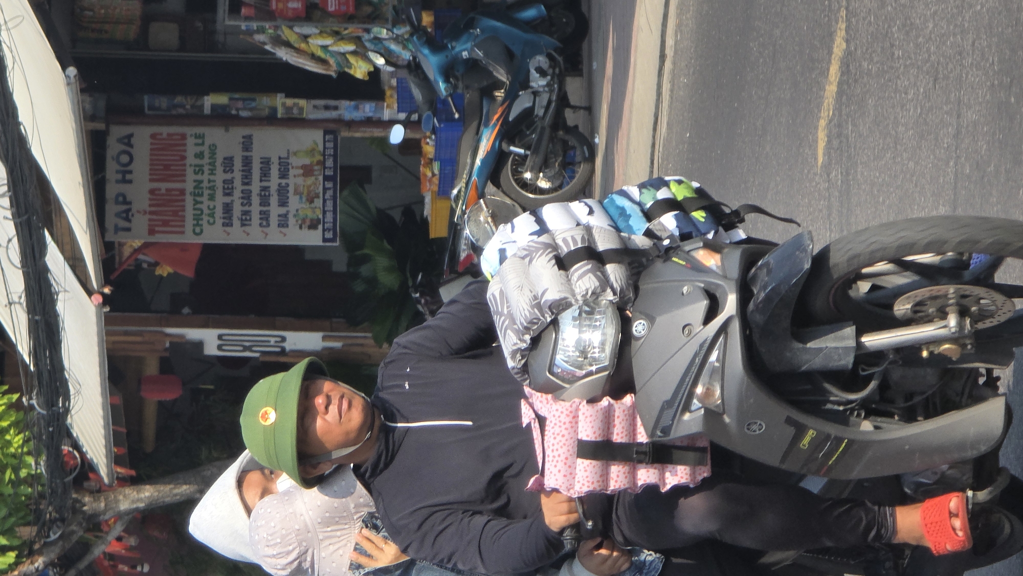 A daytime scene shows a person wearing a green cap with a red star emblem and black clothing driving a gray motorcycle on a road, as seen from a slightly elevated perspective. The motorcycle has items strapped to it. To the left, a woman with a white dotted scarf is partially visible. In the background, there is a storefront with a sign that reads "TẠP HÓA THẮNG NHUNG," offering various items such as cakes, candies, bird nests, mobile electric cars, beer, and soft drinks. Another motorcycle is parked near the storefront. The store is located in Bắc Nha Trang, Vietnam.