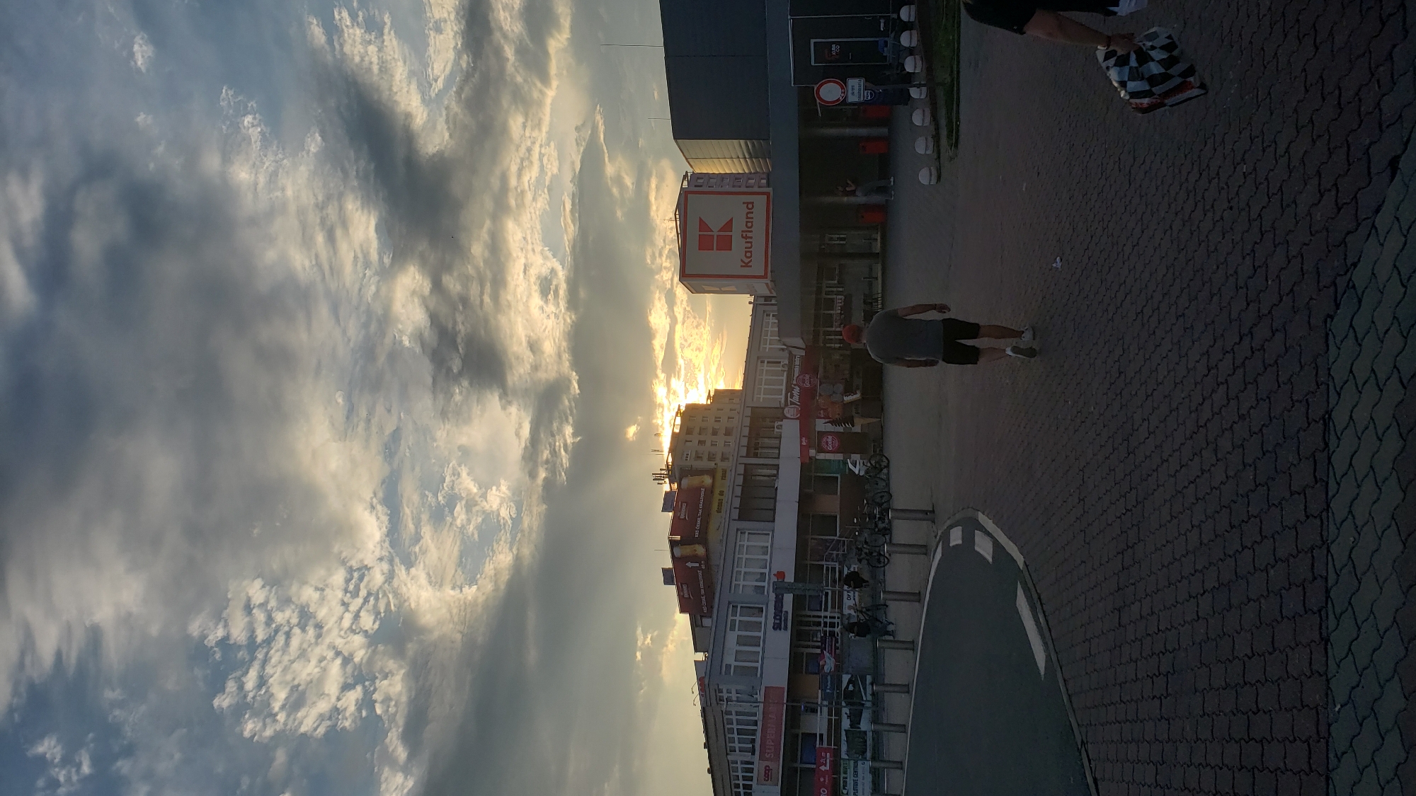 The image depicts a street scene in Prešov, Slovakia, with a prominent "Kaufland" supermarket sign visible. The sky is filled with dramatic clouds, with the sun setting or rising, casting a warm glow. In the foreground, a man in a grey t-shirt and black shorts walks away from the camera. Further back, several bicycles are parked. The architecture suggests a commercial area, likely near the Kaufland supermarket. The ground is paved with interlocking bricks.