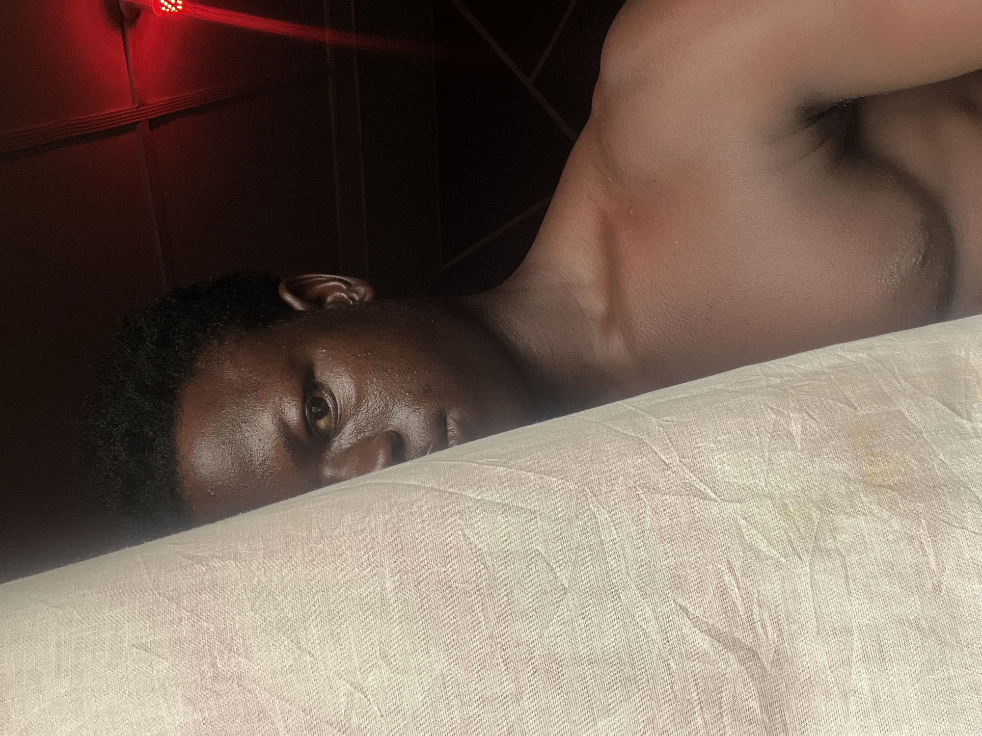 A close-up, horizontal shot depicts a dark-skinned person lying on a light-colored, wrinkled fabric, possibly a bedsheet. The person's head is positioned towards the left side of the frame, with their face partially visible. Their eye is open, and skin appears to be damp. The person's right shoulder and upper chest are visible in the upper part of the frame. The background is dark, with what appears to be a red light fixture on the upper left. The location, Mosafejaw, Nigeria is not discernable within the contents of the photograph.