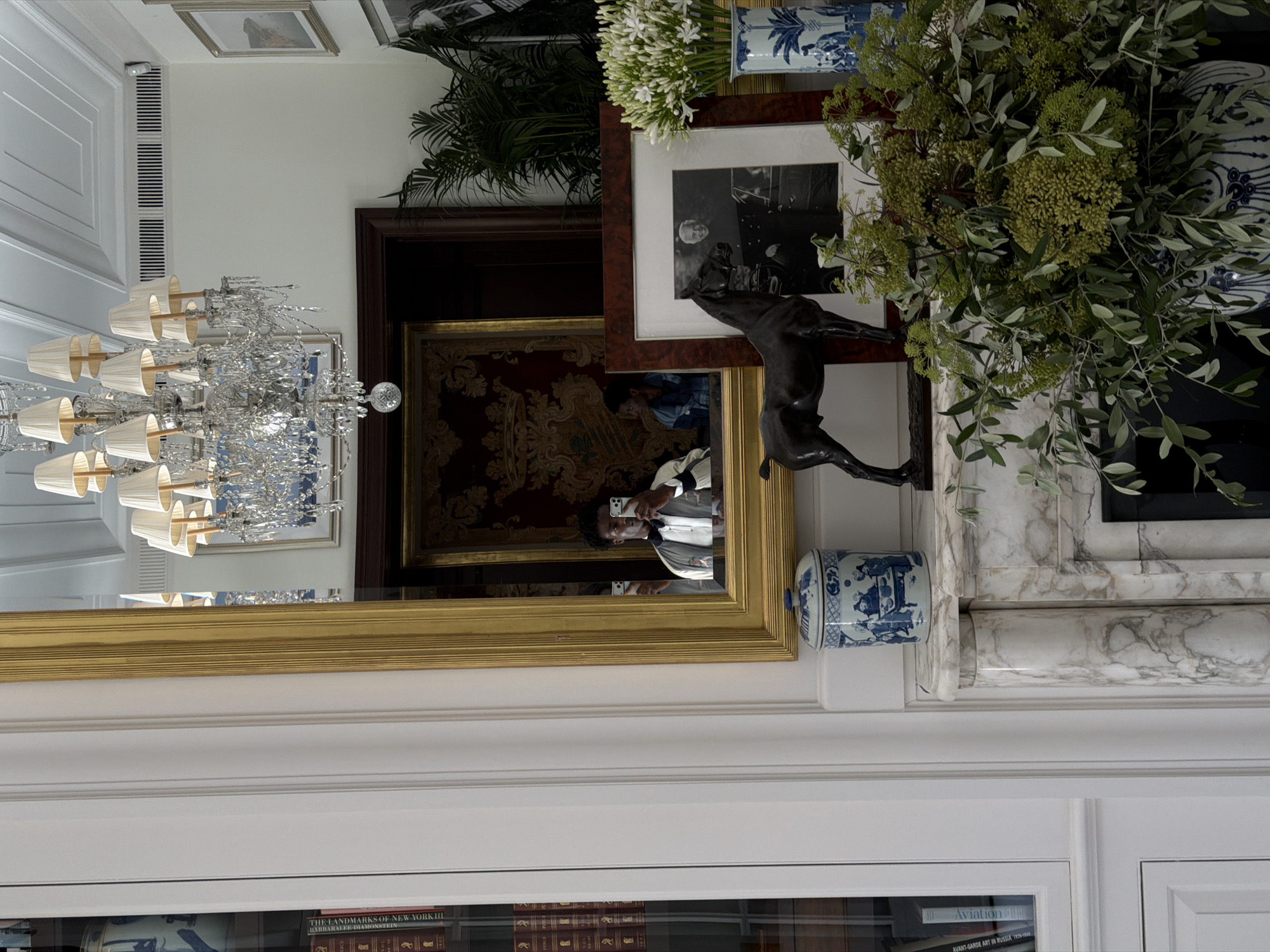 The image depicts an interior scene, possibly a residential or commercial space, with a focus on decorative objects and architectural details. A man is visible in a mirror, taking a selfie with his phone. He is wearing a formal jacket over a shirt and bow tie. On a marble fireplace mantel, a blue and white porcelain jar sits next to a dark, stylized sculpture of a horse. Behind the horse, a framed black and white photograph shows a man in uniform. A large bouquet of greenery and white flowers is placed in a blue and white vase to the right. A chandelier with multiple lampshades hangs from the ceiling. Books are visible on shelves to the left of the frame. The setting suggests a sophisticated and classic aesthetic. The city of Milan, Italy is not identifiable from the provided image.