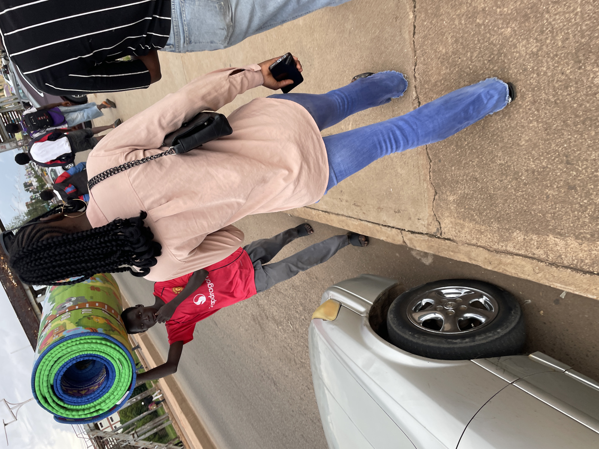 A young Black male walks along a paved sidewalk next to a road in Jos, Nigeria. He is carrying a large, rolled-up mat on his head and wears a red t-shirt with the Snapdragon logo and gray pants. To his right, a Black female walks away from the camera, wearing a pale pink button-down shirt and blue bell-bottom jeans, carrying a black purse on her shoulder and a smartphone in her right hand. Further behind them, other pedestrians are visible walking in the same direction. A silver car is parked on the side of the road, partially obstructing the view. The environment appears to be an urban setting with some overhead structures and a cloudy sky.