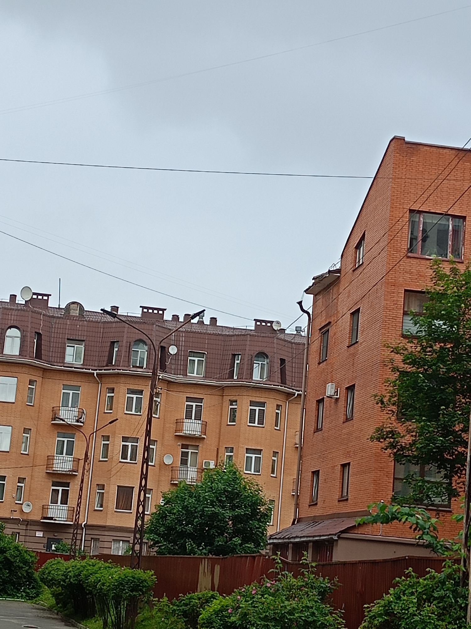 This media file captures an outdoor scene featuring two buildings and lush greenery. The building on the left is an orange, multi-story structure with a brown, mansard-style roof. It has numerous windows, some with balconies, and satellite dishes on the roof. The building on the right is a taller, brick structure with a flat roof and several windows. Both buildings appear to be residential or office buildings.
The scene is set in a slightly overcast environment, suggesting it might be daytime with cloudy weather. The presence of trees and bushes indicates an urban or suburban setting with landscaping. The buildings are close to each other, and electrical wires are visible crisscrossing the sky.
There is no visible text in the image, and no people are present. The overall impression is a quiet, possibly residential, area on a cloudy day. The composition focuses on the architecture of the buildings and the surrounding vegetation. The lighting suggests it is not dawn or dusk. The condition of the buildings suggests they are in use, with satellite dishes and air conditioning units visible on the brick building.