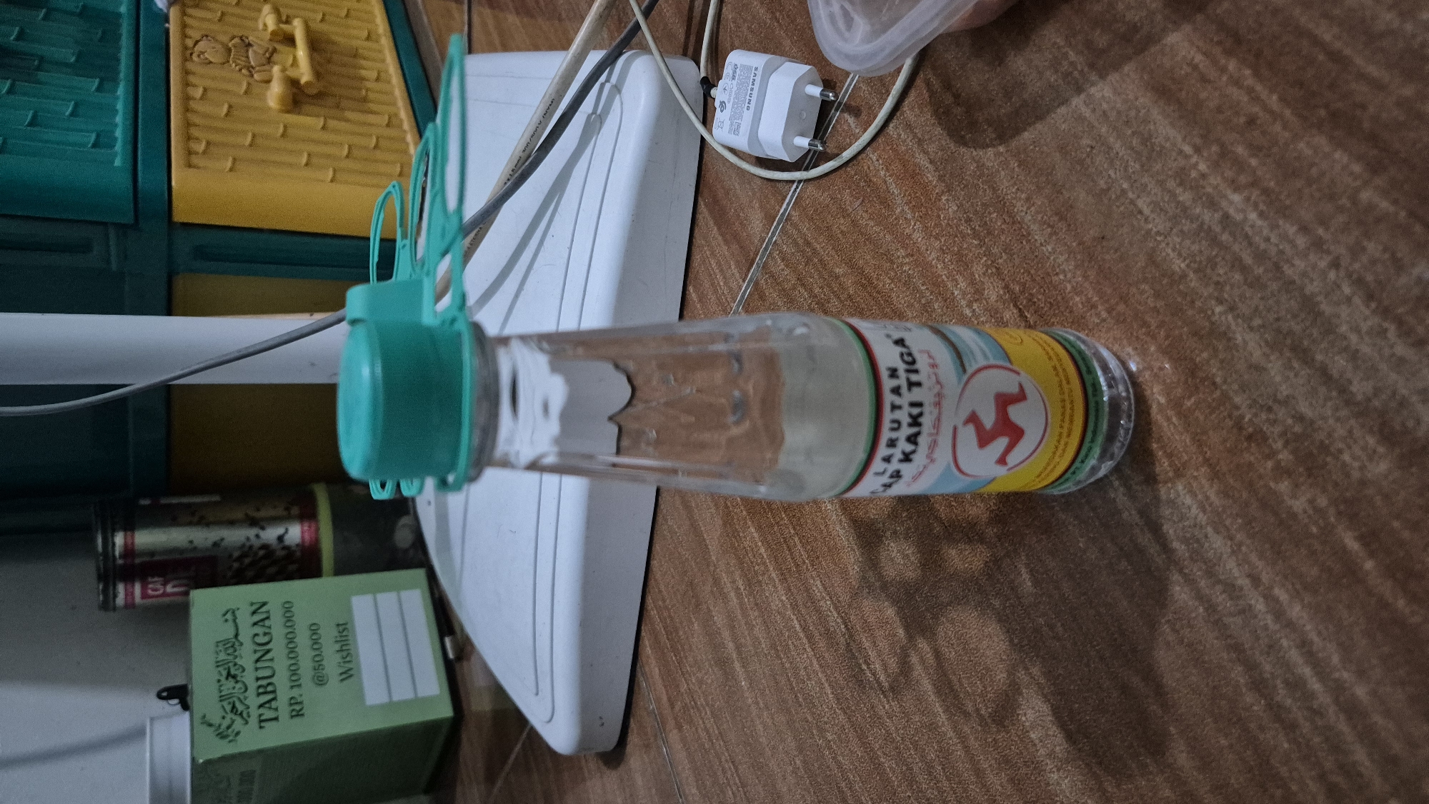The image depicts an indoor scene on a brown, wood-patterned floor. In the foreground, a clear glass bottle with a teal-colored cap and an attached plastic tamper-evident seal is centrally positioned. The bottle contains a clear liquid with some settled sediment at the bottom. A label on the bottle reads "LARUTAN KAKI TIGA" (Three-Legged Brand Solution), with some Arabic script above it and a circular red and yellow logo featuring a stylized red figure.
Behind the bottle, a white, flat appliance, possibly a fan base or a humidifier, is visible. To the left of the bottle, a green box with Arabic script and the word "TABUNGAN" (Savings) printed on it is present. The box also indicates "RP. 100.000.000" and "@50.000 Wishlist." Further in the background, there are two cylindrical metal cans, one with a red label. To the right of the white appliance, stacked plastic storage containers in green and yellow are visible, with a teddy bear design on the yellow one. A white Samsung power adapter with two prongs is plugged into an unseen outlet, and its white cable extends towards the bottom right of the frame. The lighting suggests an indoor setting, possibly a home. The image does not provide any specific geographical markers to indicate a location in the city of Karawang, Indonesia.