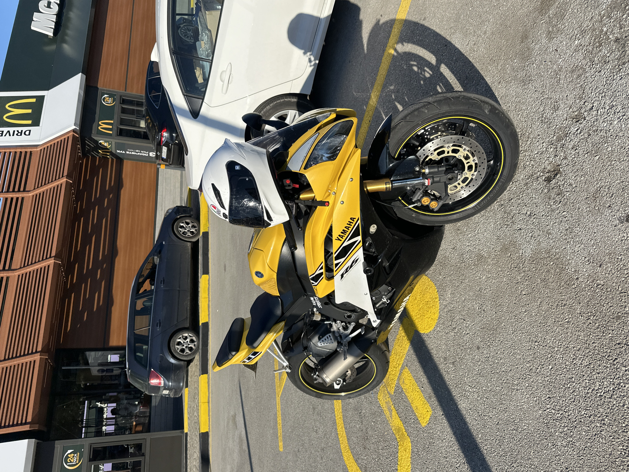 The image shows a yellow Yamaha R6 motorcycle parked outside a McDonald's restaurant. The motorcycle is in the foreground, with a white car and a dark-colored car parked behind it. The McDonald's building is visible in the background, with its distinctive golden arches logo. The scene is set on a sunny day, with shadows cast on the asphalt. Yellow and black painted lines on the parking lot indicate designated parking spots and traffic flow.
Visible text includes "DRIVE THRU" on the McDonald's signage. The motorcycle has "YAMAHA" branding. There is also a "24 HOUR" sign with a clock icon on another part of the building.
The setting appears to be a typical fast-food drive-thru in Veliko Tarnovo, Bulgaria. The bright sunlight suggests it is daytime. The overall mood is ordinary, depicting a common urban scene. There are no people prominently featured in the image, but some figures can be seen through the windows of the dark car and the restaurant. The focus is on the parked vehicles and the McDonald's establishment.