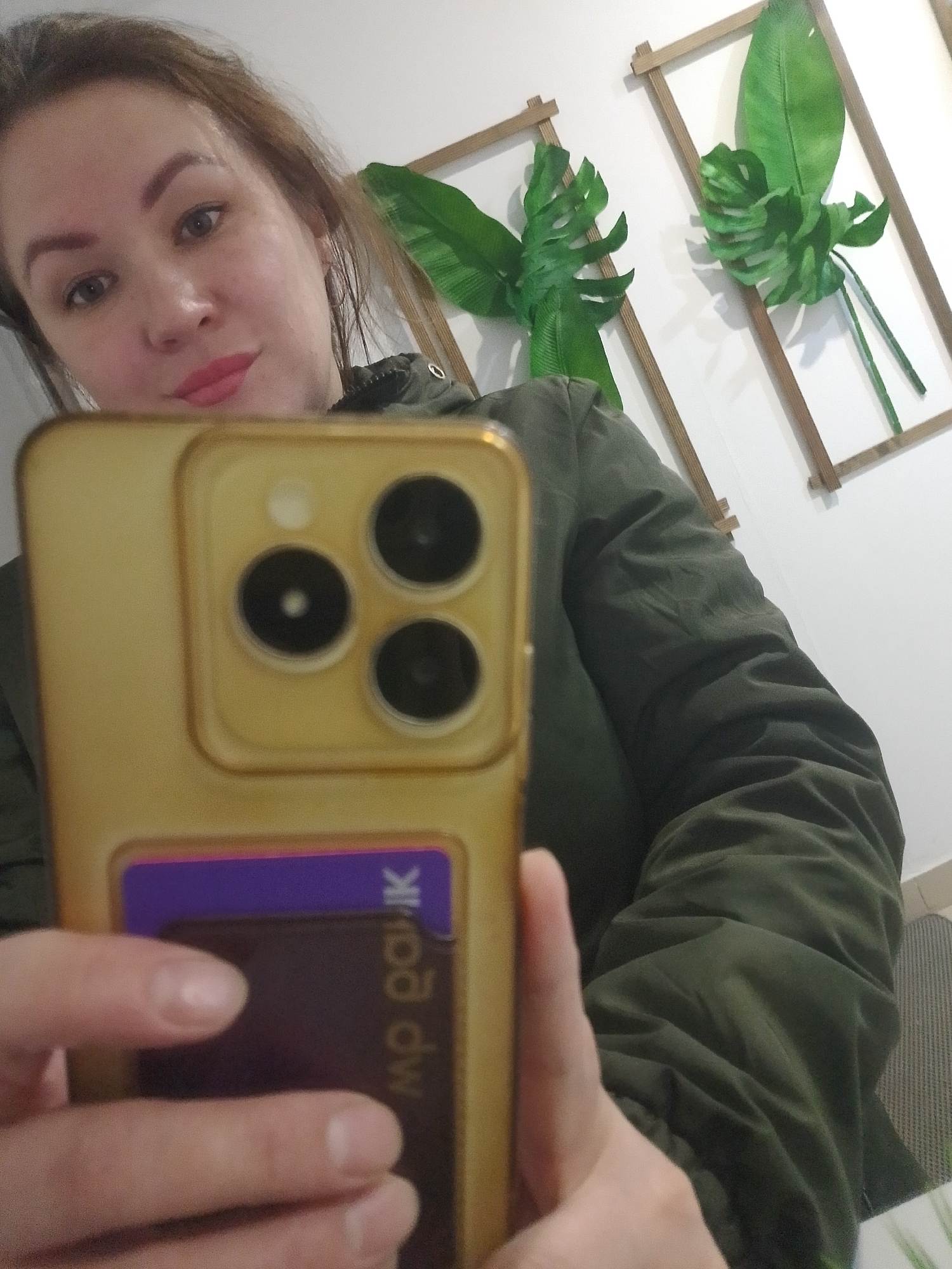 A woman takes a mirror selfie indoors in Chaikovsky, Russia. She is partially visible, focusing on her face and upper body, holding a golden-colored smartphone with her right hand. The phone features a prominent triple-camera module on its back, typical of modern high-end smartphones. A purple and dark brown card is tucked into the back of the phone case, displaying partially visible, mirrored text that appears to read "mp paik" or similar.
The woman has light skin, dark brown hair, and wears a dark green, possibly olive-green, quilted jacket. Her lips are painted a coral or light pink shade, and she looks directly towards the camera with a neutral to slightly pleasant expression. Her fingernails are short and unpainted.
In the background, behind her, two decorative wall hangings are visible on a white wall. Each hanging consists of a light wooden rectangular frame enclosing large artificial green leaves, resembling monstera and banana leaves, arranged as tropical wall art. The lighting suggests an indoor setting, and no specific time of day or weather is discernible.
