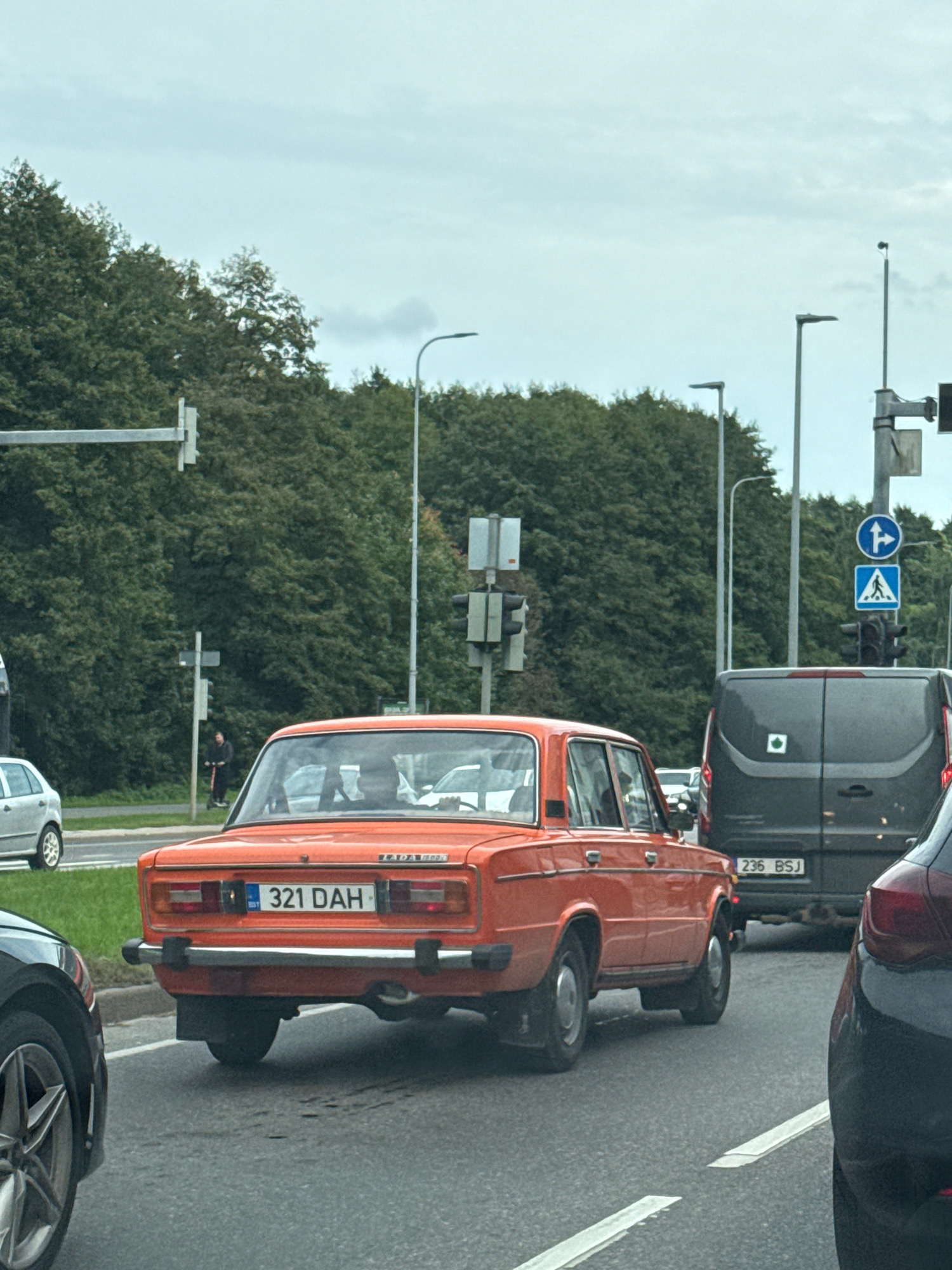 The image depicts a street scene in Tallinn, Estonia. In the foreground, an orange Lada car with license plate "321 DAH" is visible. To the right of the Lada, a dark-colored van with license plate "236 BSJ" is present. Further to the right, the rear of a dark-colored car is partially visible. In the background, a row of trees lines the street, and traffic lights and streetlights are observed. A person is seen standing on the sidewalk, operating an electric scooter. The sky is overcast.