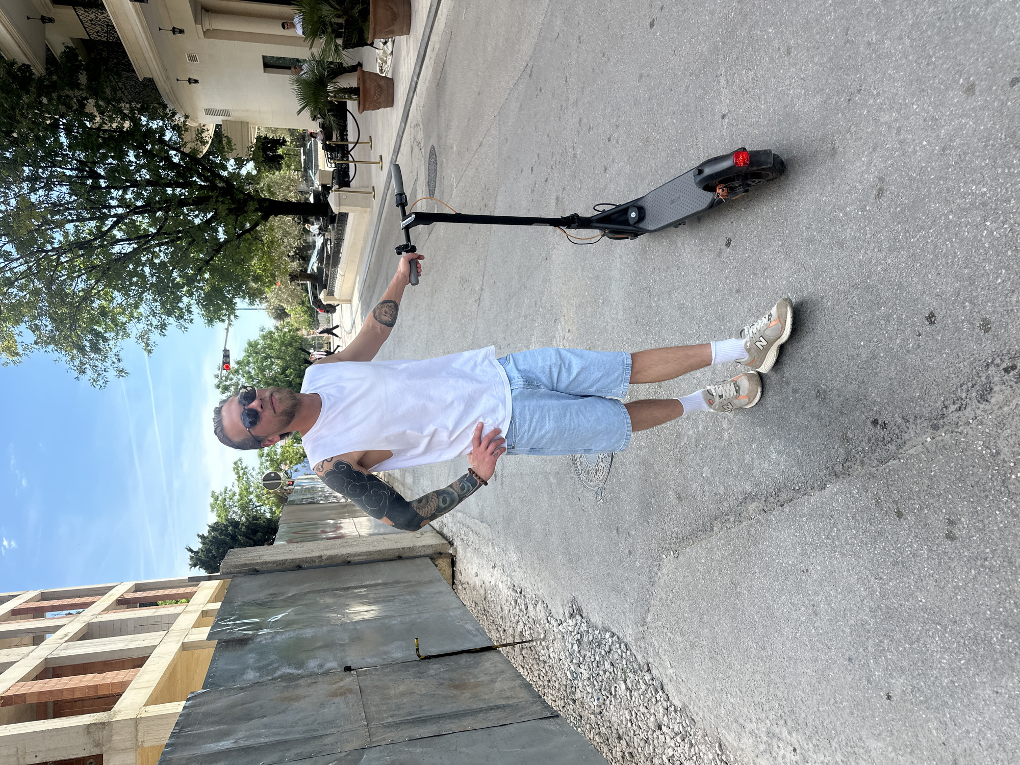 A man with a shaved head, sunglasses, and extensive tattoos is standing on a sidewalk in Budva, Montenegro, next to an electric scooter. He is wearing a white tank top, light blue shorts, white socks, and light-colored sneakers. The man is posing with one arm raised and the other on his hip.
The scooter is black and gray and is lying flat on the sidewalk. Behind the man, there is a metal barrier and an unfinished concrete building. In the background, there is a street with cars, trees, and buildings. The sky is blue with some clouds.