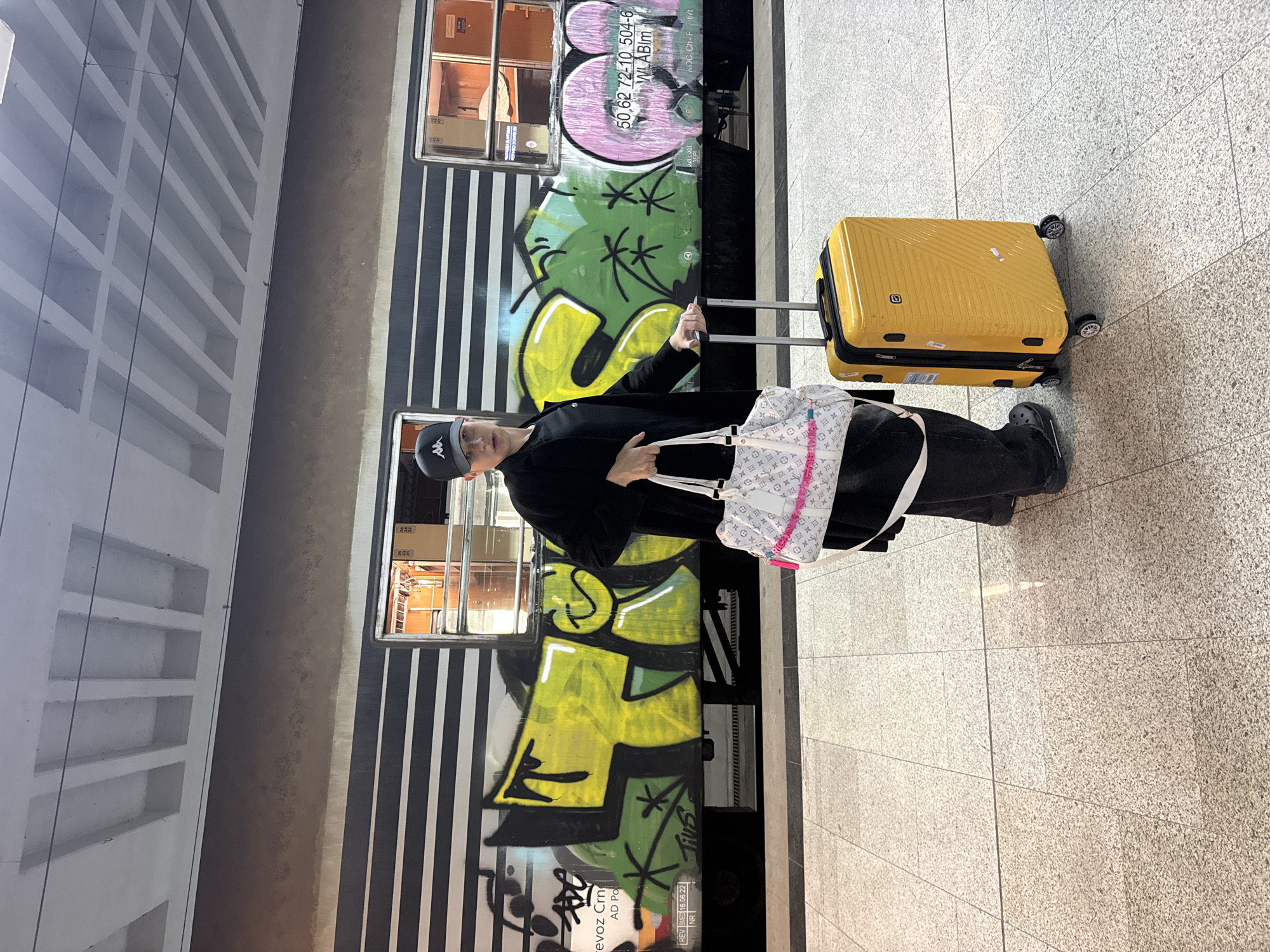 A person wearing a black Kappa baseball cap, a black jacket, and black pants stands next to a train car with graffiti, holding the handle of a yellow suitcase. The person also has a white purse with pink accents slung over their shoulder. The train car features vertical black and white stripes and graffiti. The text on the train car includes "revoz Crn AD Po," "REV SMED 16.06.22 NR," "Tius," "50.62 72-10 504-6 WLABIM," and "LOC.Ch-F 6/1." The location is characterized by tile flooring and is likely a train station. Based on the text "revoz Crn," it is possible that the location is in the city of Belgrade, Serbia.