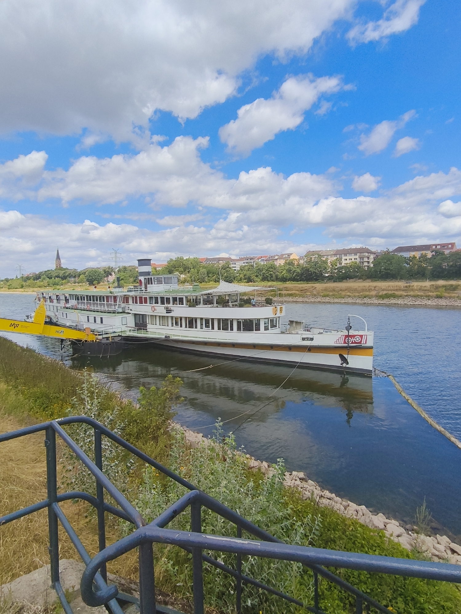 A white paddle steamer with yellow accents is docked on a wide, blue river. The steamer has multiple decks with numerous windows and a tall smokestack. A yellow ramp or gangway is attached to the side of the boat. The riverbank on the right is lined with large rocks and green vegetation, while the riverbank on the left is covered in dry grass and more vegetation. In the background, across the river, is a city skyline with buildings and trees. A church steeple is visible to the left of the city. The sky is a vibrant blue with scattered white clouds. A dark blue metal railing is in the foreground on the left, descending towards the river. The scene appears to be in Mannheim, Germany, likely on the Rhine River. There are no visible people in the image.