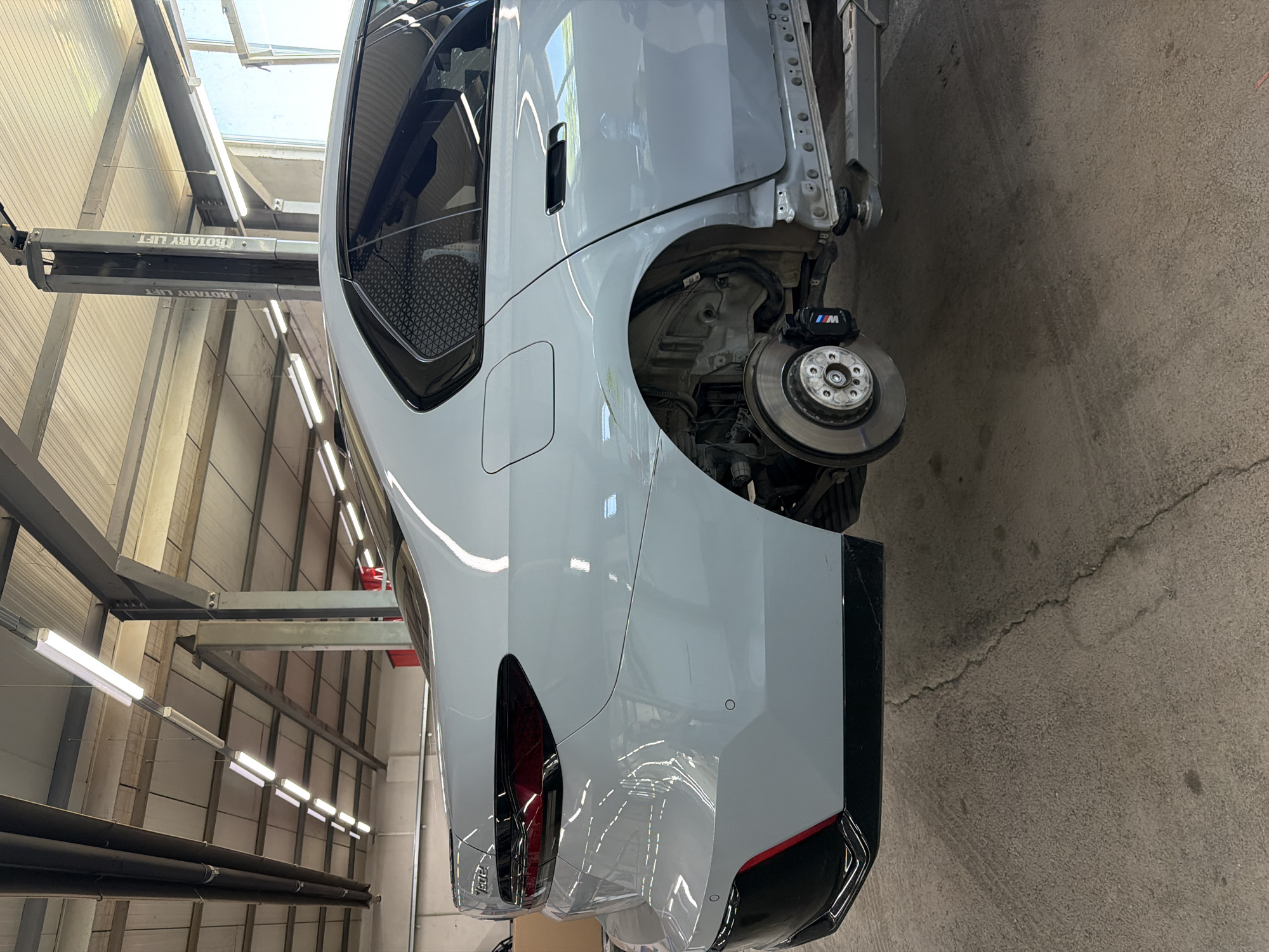 The image shows a white car being worked on in a garage. The car is lifted on a rotary lift, and the rear wheel is removed, exposing the brake caliper and rotor. The car appears to be undergoing some form of repair or maintenance. The garage has industrial lighting and a concrete floor. The overall scene suggests a professional auto repair setting. The image is oriented vertically, and the car is viewed from the rear, three-quarters perspective.
Notable details include the text "ROTARY LIFT" visible on the lift mechanism. The brake caliper has the "M" logo, likely indicating it's part of a performance package. The car has a sleek, modern design with dark-tinted taillights. The setting is indoors, likely a workshop or garage, with bright fluorescent lighting. No people are visible in the image. The environment appears clean and organized, typical of a professional automotive service center. Given the location context of Burgas, Bulgaria, the image captures a moment in an automotive workshop in that region.