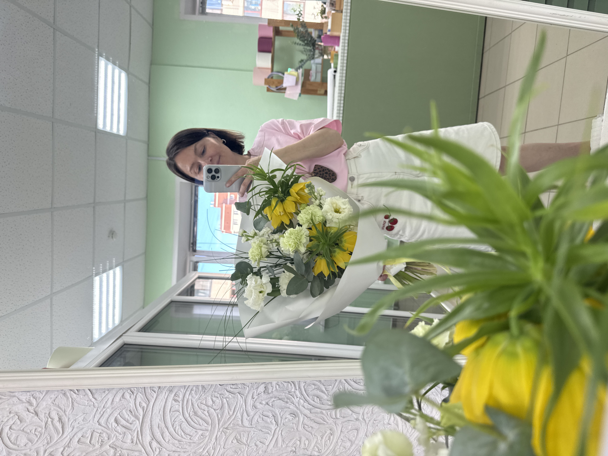 A person is reflected in a mirror, holding a bouquet of yellow and white flowers and a phone. The person has dark hair and is wearing a pink shirt and white shorts with cherries on them. The background includes a green wall, a window, and a white ceiling with fluorescent lights. The mirror has a decorative frame. The location could be inferred as a shop or store due to the arrangement and nature of the background elements. It is unknown if it is in Izhevsk, Russia.