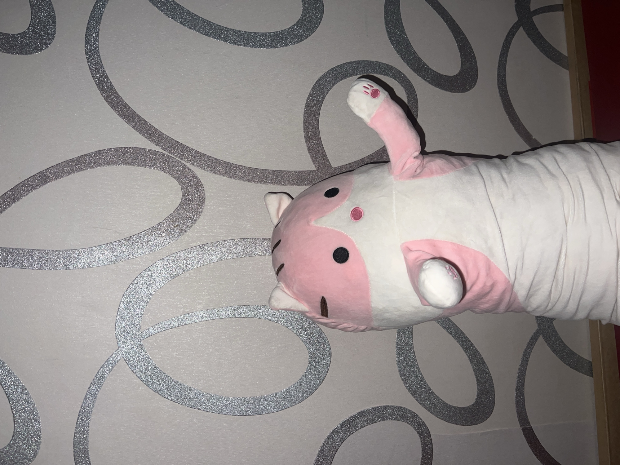 A large, plush, white and pink animal-shaped pillow or body pillow is depicted lying against a wall. The plushie, which resembles a stylized cat or similar creature, features a pink face with black oval eyes, a small pink nose, and stitched mouth details. One pink paw with visible pink paw pads is raised. The body of the plushie is long and cylindrical, covered in white, somewhat wrinkled fabric, suggesting it is a pillow or soft toy.
The background is a wall covered in light gray or off-white wallpaper. This wallpaper is adorned with prominent, glittery silver-gray abstract swirling and looping patterns. On the right edge of the frame, a thin strip of light wood and a glimpse of a dark red surface are visible, possibly part of a door frame or piece of furniture.
The scene is an indoor setting, likely a room. The lighting appears artificial. No human activity is taking place; the plushie is simply resting or displayed against the wall. There is no visible text in the image. The location context for this scene is Kazan, Russia.