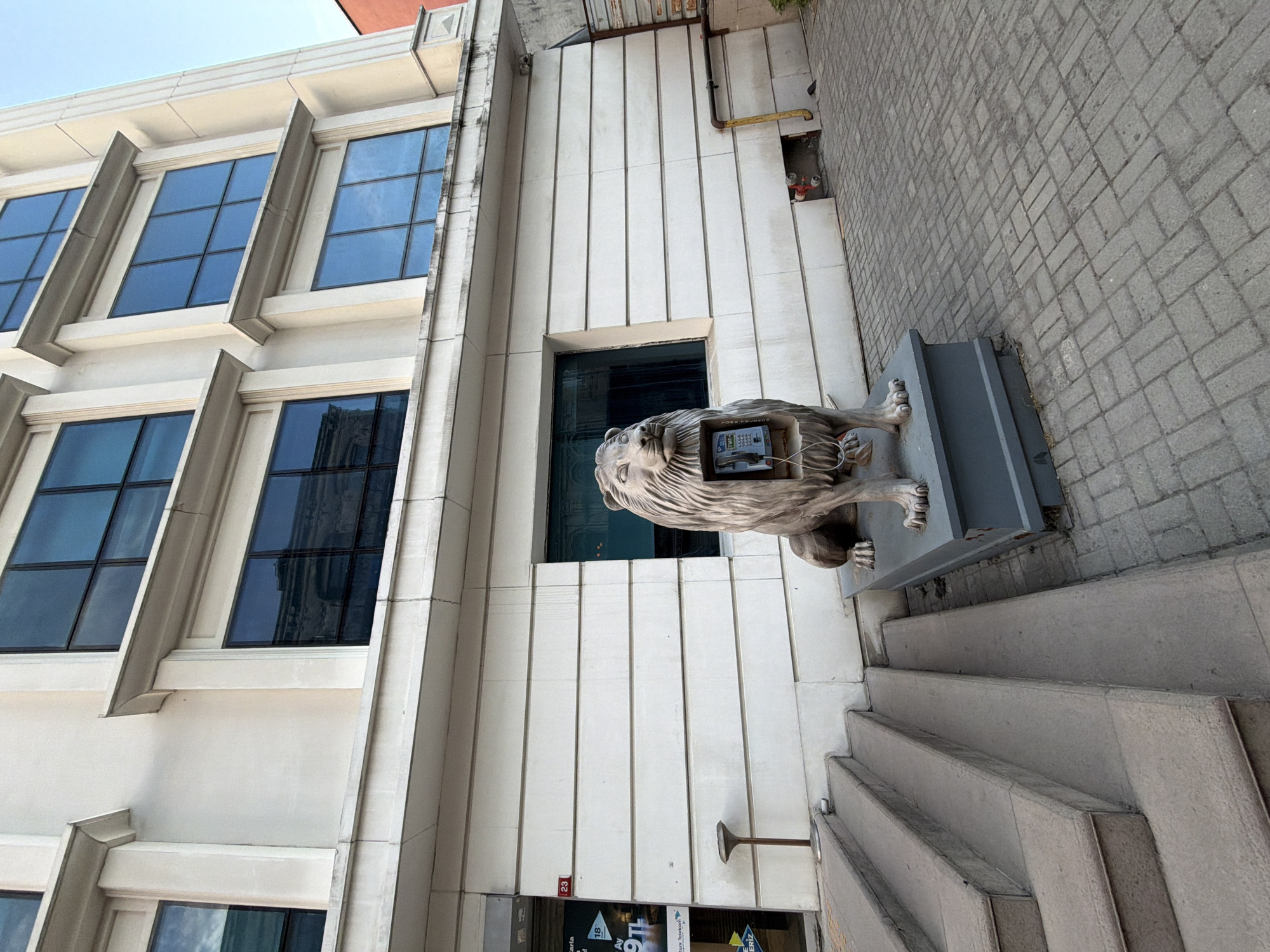 This is a full shot of a lion statue with a phone booth integrated into its chest, located on a city street in Istanbul, Turkey. The statue is made of concrete and painted a stone gray. It is positioned in front of a building with white facade and large blue-tinted windows. The building appears to be an office building or a commercial establishment. To the left of the statue, there is a set of concrete stairs leading up to an entrance. A small, brown lamp post stands on the landing of the stairs. The ground is paved with gray bricks. The statue is the focal point of the image, with the phone booth inside its chest being clearly visible. The sky is clear and blue, suggesting it is daytime. The overall scene is static and captures an unusual urban art installation.