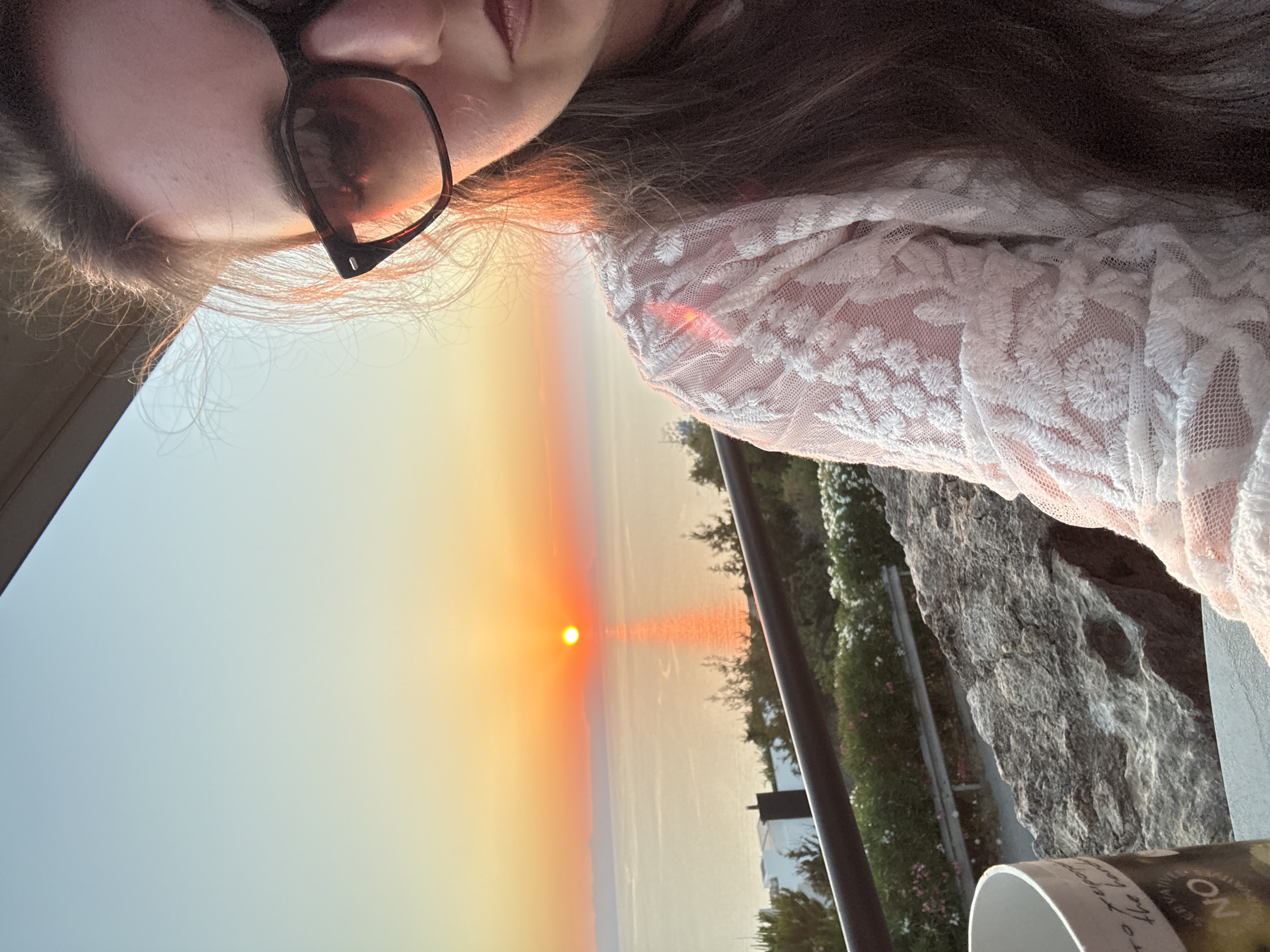 A selfie shows a woman with long, dark hair and dark-rimmed sunglasses. She is wearing a white lace top. The background is a sunset over the ocean, with the sun appearing as a bright orange orb on the horizon. The sky transitions from a pale blue at the top to a warm orange near the horizon. In the foreground, there is a rocky ledge and some greenery. A white mug is partially visible on the left side of the image. The scene appears to be outdoors, possibly on a balcony or terrace overlooking the sea.