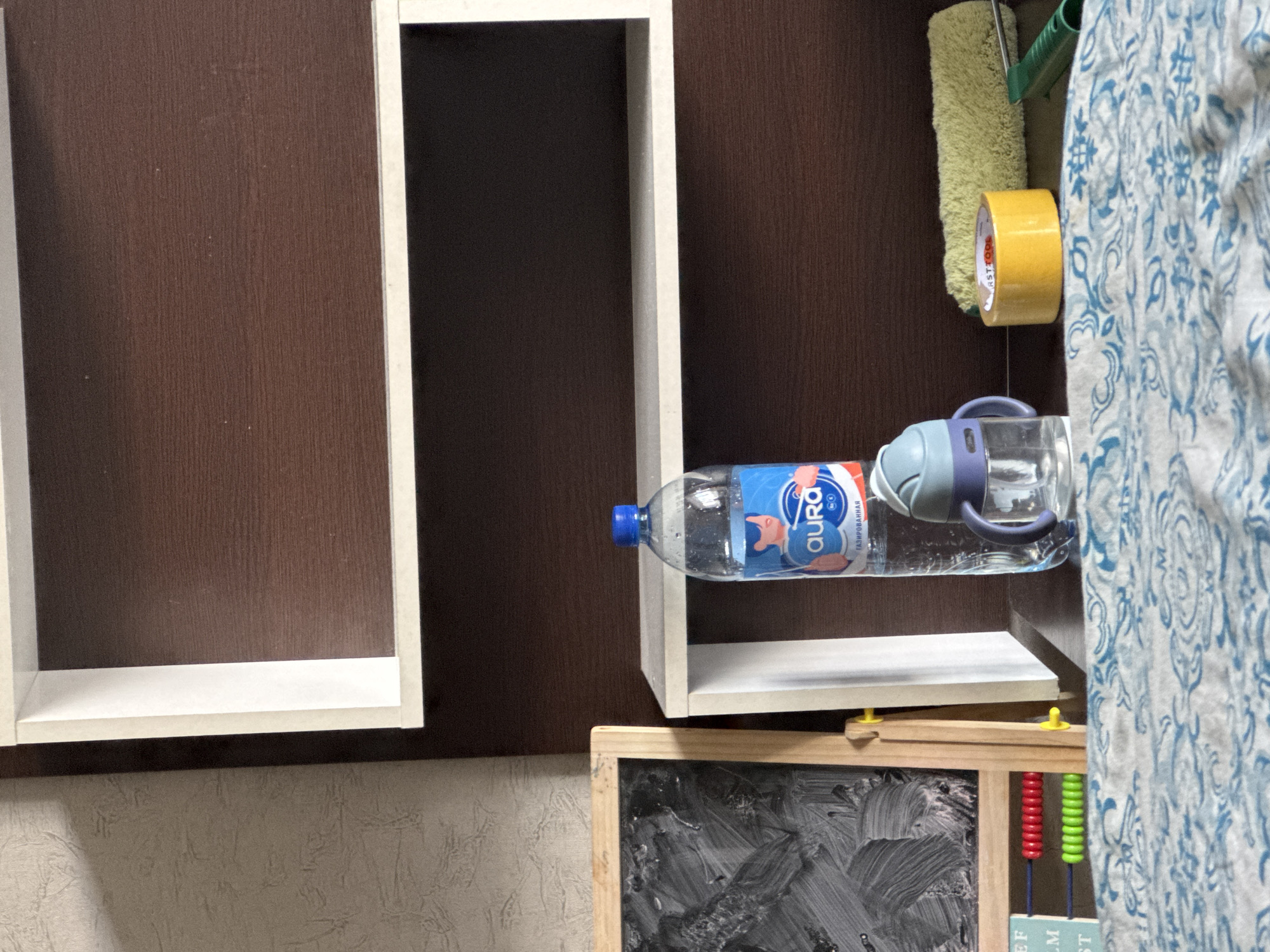 The image depicts an indoor scene, likely a room, with a focus on a wooden shelf unit against a textured wall. On the bottom shelf, several objects are arranged. To the left, a small blackboard with a wooden frame stands on an easel, showing smudged grey paint. Below the blackboard, an abacus with red and green beads is visible, along with a portion of a blue and white patterned fabric in the foreground. In the center of the shelf, a clear plastic bottle of Aura mineral water with a blue cap stands upright. Next to the water bottle is a small, clear sippy cup with a blue lid and handle. To the right of the sippy cup, a roll of yellow tape sits on the shelf, and beyond that, a fuzzy green object, possibly a cleaning tool, is positioned next to a green plastic handle. The overall impression is of a workspace or a child's area. The location is not specified in the image.