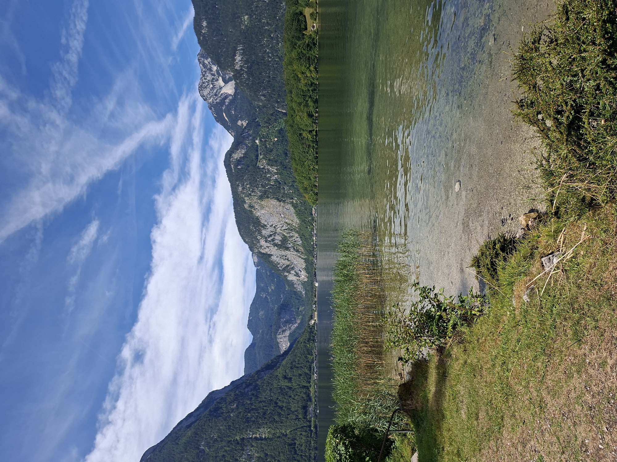The image shows a panoramic view of Hallstätter See, a lake in Obertraun, Austria. The lake is calm and clear, with the bottom visible in some areas. The foreground features a grassy bank with some reeds and small bushes. In the background, towering green mountains rise on either side of the lake, their slopes covered in dense forest. The sky is a vibrant blue with wispy white clouds. On the far side of the lake, a small village with red-roofed houses is nestled along the shore, with more mountains in the distance. The scene is idyllic and tranquil, evoking a sense of natural beauty and peace.