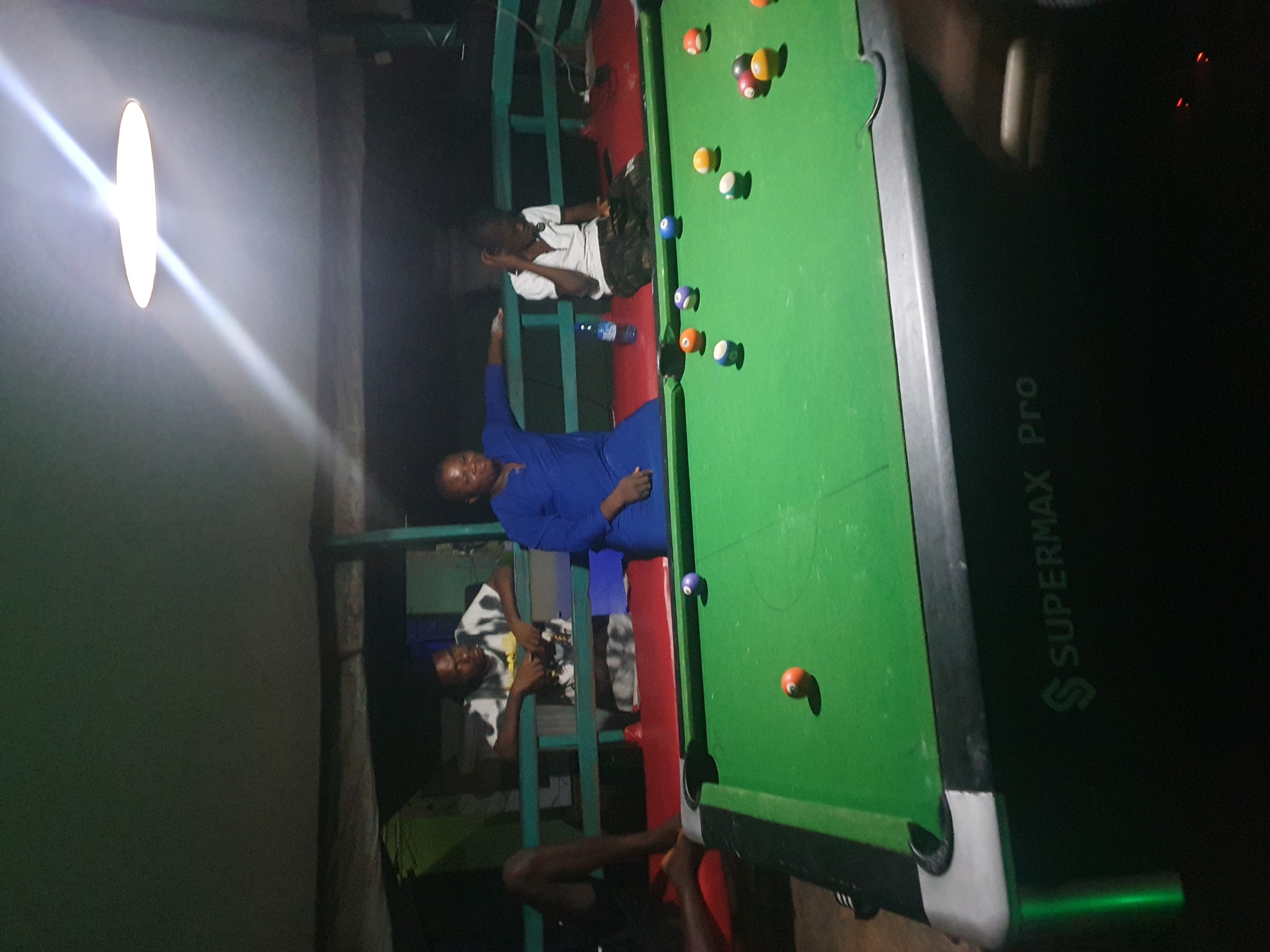 In Lokoja, Nigeria, a group of individuals gathers around a pool table located outdoors under a covered structure. The lighting is dim, primarily illuminated by a circular light fixture overhead, casting a warm glow and strong reflections on the ceiling.
In the foreground, a green pool table dominates the scene, with various colored balls scattered across its surface. One orange ball is positioned near the middle of the table, and a blue ball is closer to the top edge. The table itself is branded "SUPERMAX Pro" on its side.
Three men and one woman are visible. To the left, a man is seated with his legs crossed, facing away from the camera. To his right, a man in a tie-dye t-shirt leans over the railing of the structure, looking towards the pool table. A woman in a vibrant blue dress is seated next to him, also looking towards the table. To the far right, a young boy in a white t-shirt sits with his hand to his head, appearing engaged with the game. A blue water bottle rests on the red surface behind the pool table. The environment suggests a relaxed, recreational setting, possibly a backyard or a local hangout.