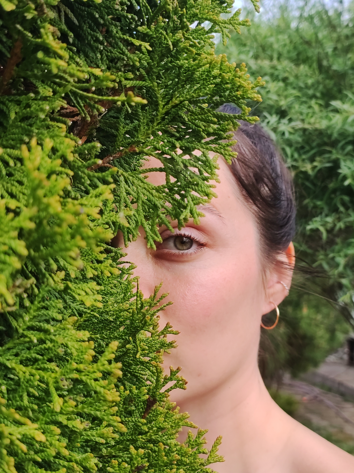 A close-up shot shows a woman partially obscured by a lush green shrub. The right half of her face is visible, revealing a green eye, fair skin, and a gold hoop earring. Her dark hair is pulled back. The dense greenery occupies the left portion of the frame, with additional green foliage visible in the background. The location is unknown and cannot be determined from the image.