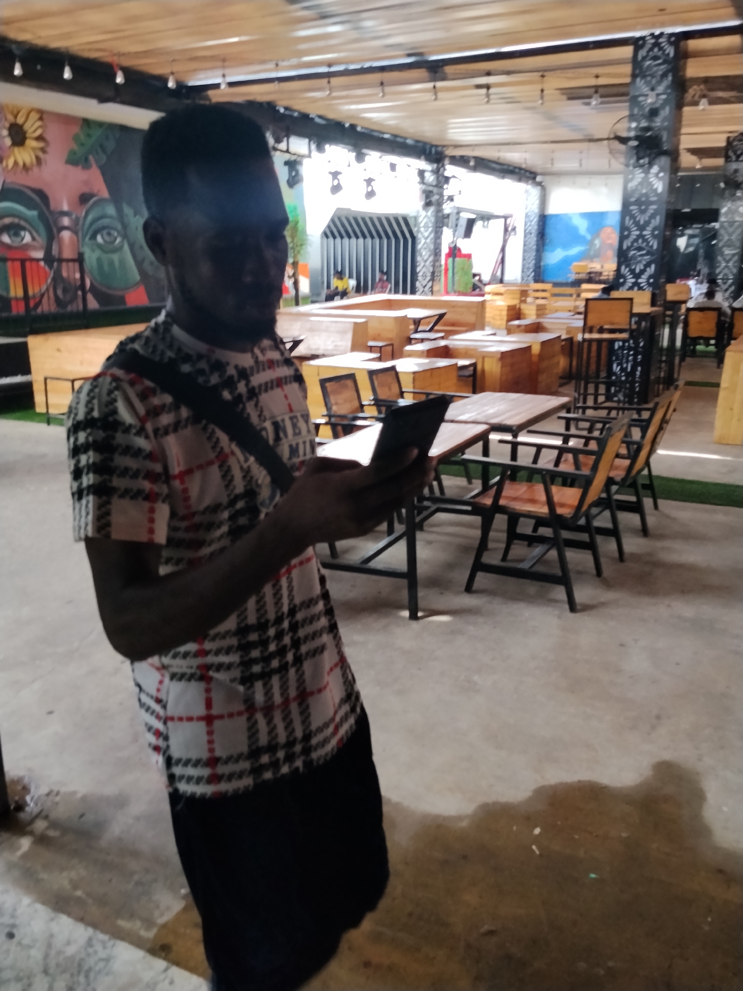 A man stands in an outdoor seating area, possibly a restaurant or bar, in Howayi, Nigeria. He is wearing a plaid shirt with black, white, and red patterns and a black crossbody bag. He holds a mobile phone in his hands, appearing to look at it. The environment is furnished with wooden tables and chairs, some with metal frames. In the background, there are murals painted on the walls, including one with large eyes and a sunflower, and another with a blue sky and a figure. The floor is concrete, with some areas appearing wet. The ceiling is made of wooden planks with visible light fixtures. The scene suggests a casual, possibly lively, gathering space within the city.