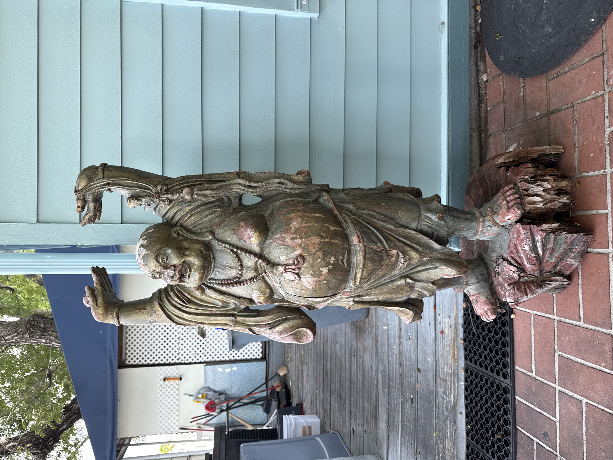 A weathered, wooden statue of a laughing Buddha is positioned outdoors on a brick walkway. The statue, which stands on a rough, carved wooden base, depicts the deity with a large belly, arms raised, and a wide smile. Its surface is worn, showing patches of reddish-pink and green discoloration beneath the predominantly golden-brown hue. The Buddha is adorned with beaded necklaces. In the background, a light blue, horizontally-paneled wall forms the backdrop. To the left of the statue, a wooden deck with gray planks is visible, featuring a black rubber mat and various cleaning tools, including mops and brooms, leaning against a white latticed structure. A large blue tarp is stretched above this area. The overall scene suggests an outdoor setting, possibly a courtyard or porch area, in Key West, Florida, United States of America.