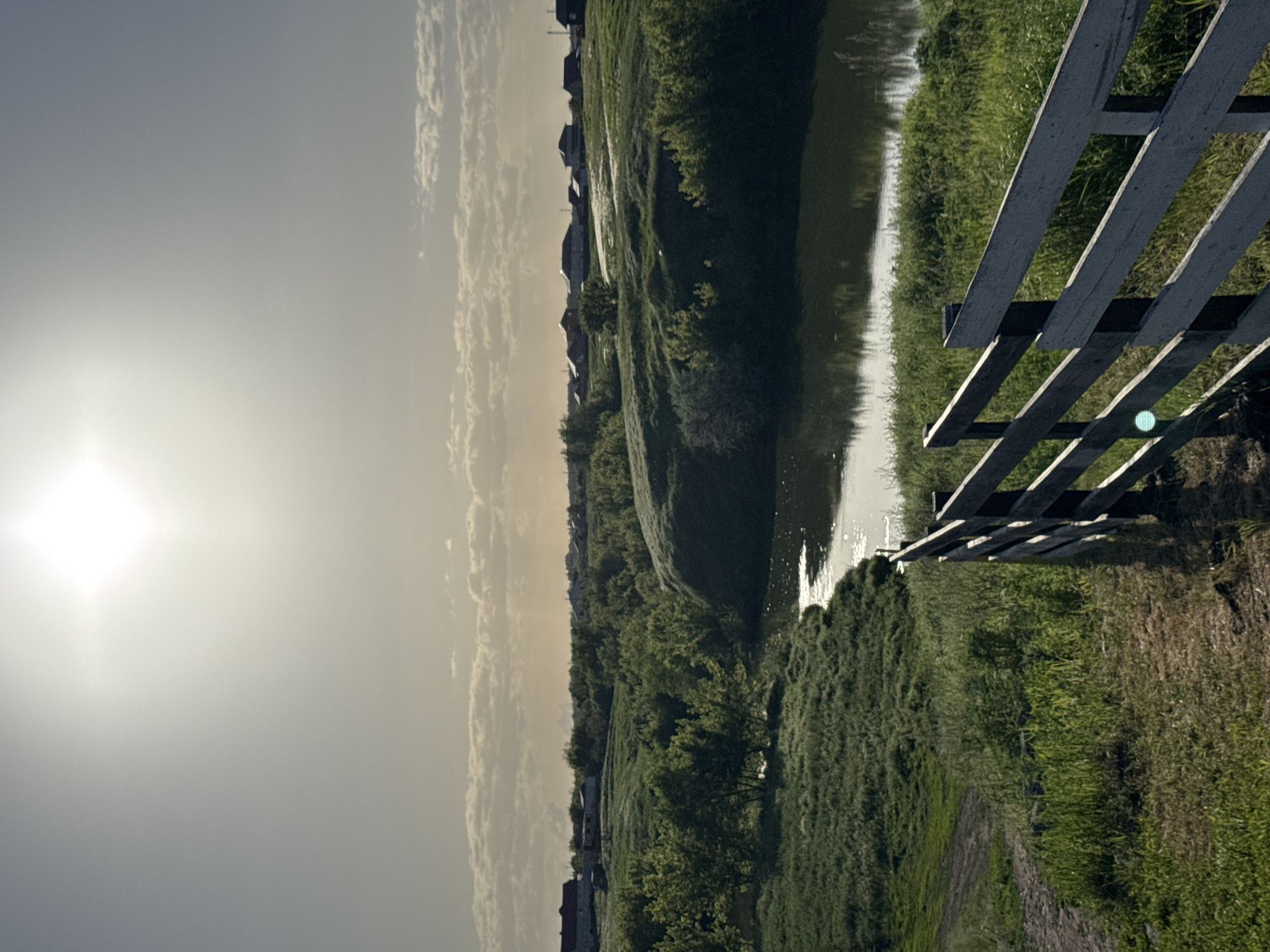 The image depicts a rural scene in Усть-Курдюм, Russia, featuring a body of water flanked by lush vegetation. A weathered wooden fence stands prominently on the right side of the frame. The sky is overcast with a bright sun visible in the upper left corner. Houses are visible in the background.