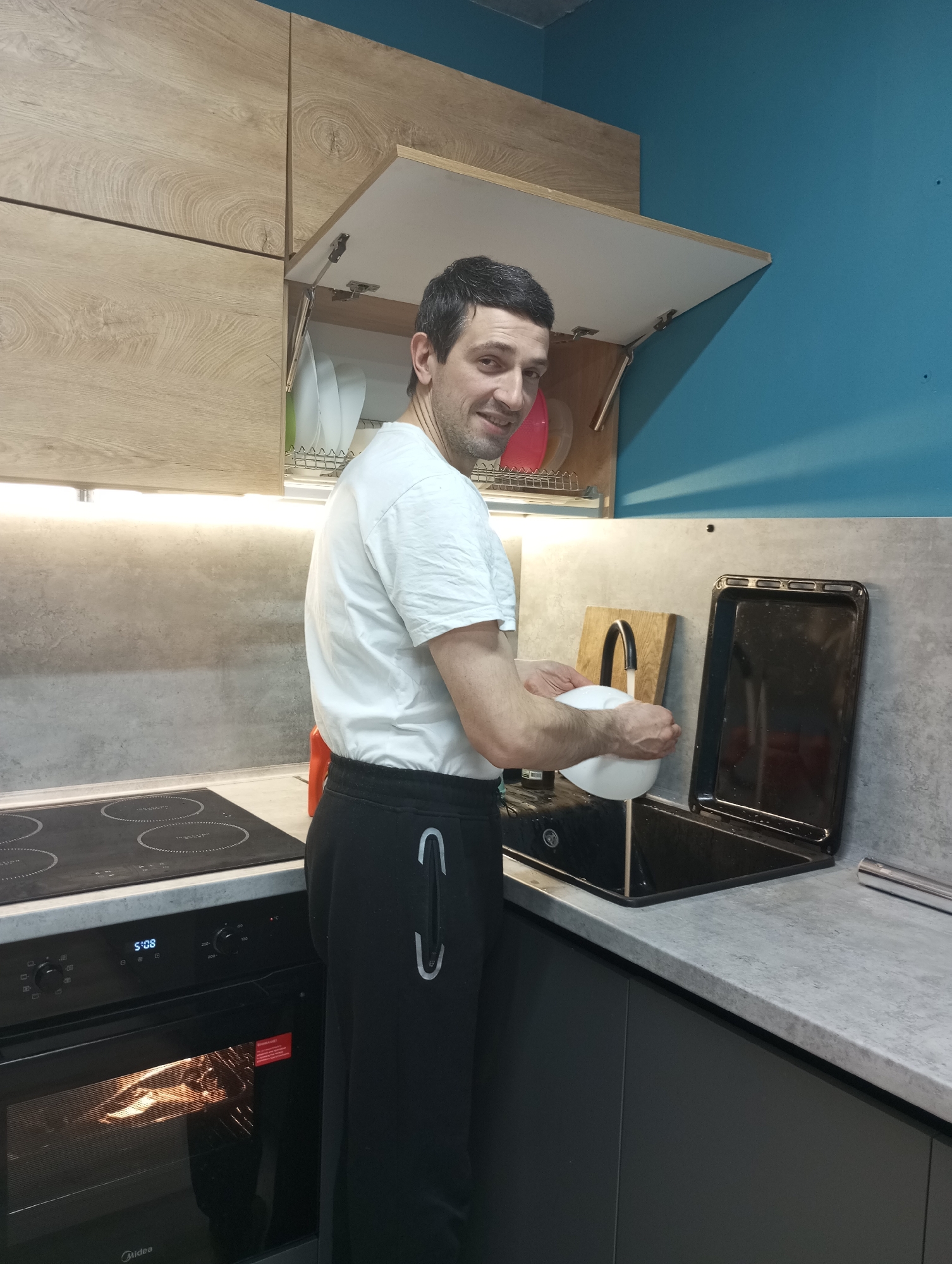 A man is in a kitchen in Pervomaysky, Russia. He is wearing a white t-shirt and black sweatpants. He is standing at a sink, holding a white bowl under a running faucet. An open cabinet is above the sink, revealing plates and a dish rack. Next to the sink is a black oven tray and a wooden cutting board. The oven below the stovetop is on, showing food inside. The kitchen has modern grey countertops and light wood cabinets. A blue wall is visible in the background.