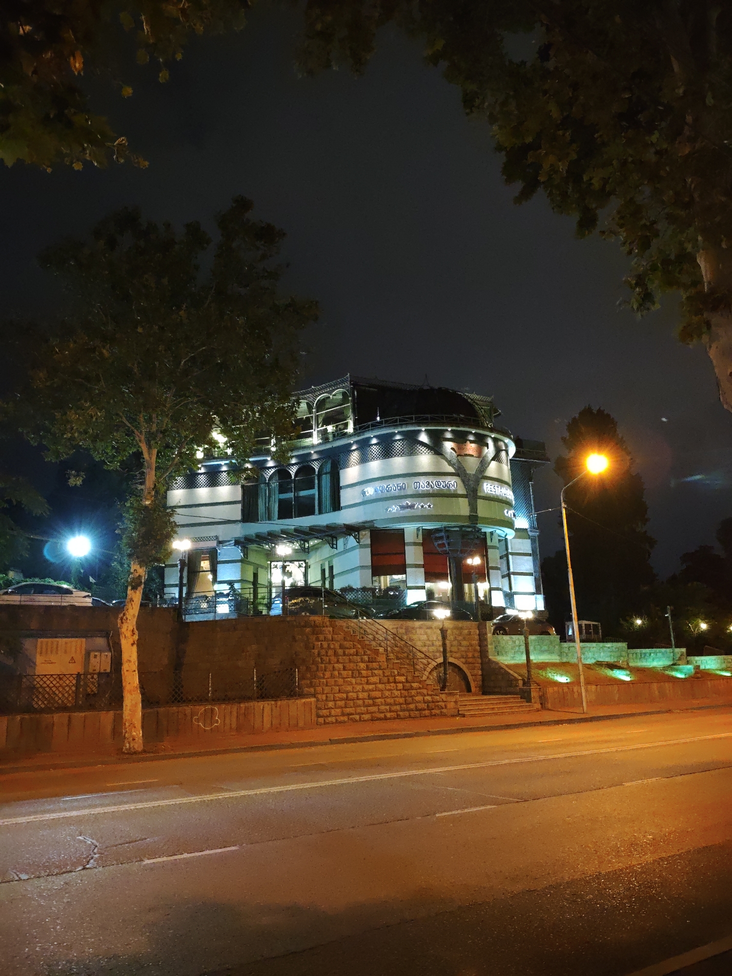 The image depicts a nighttime view of the "Chveni Galereia" restaurant in Tbilisi, Georgia. The building, characterized by its distinctive architecture with multiple levels and ornate detailing, is illuminated by external lighting. A prominent streetlamp with a bright orange glow stands to the right of the building. The foreground shows a paved road with white markings, suggesting it is a street in a city. To the left of the road, a tree with a light-colored trunk and green foliage is visible. Behind the tree and extending towards the building is a retaining wall constructed of stone. Further back, a low wall with a metal fence runs along the property line. The sky is dark, indicating nighttime. The overall scene conveys a commercial and urban setting at night.