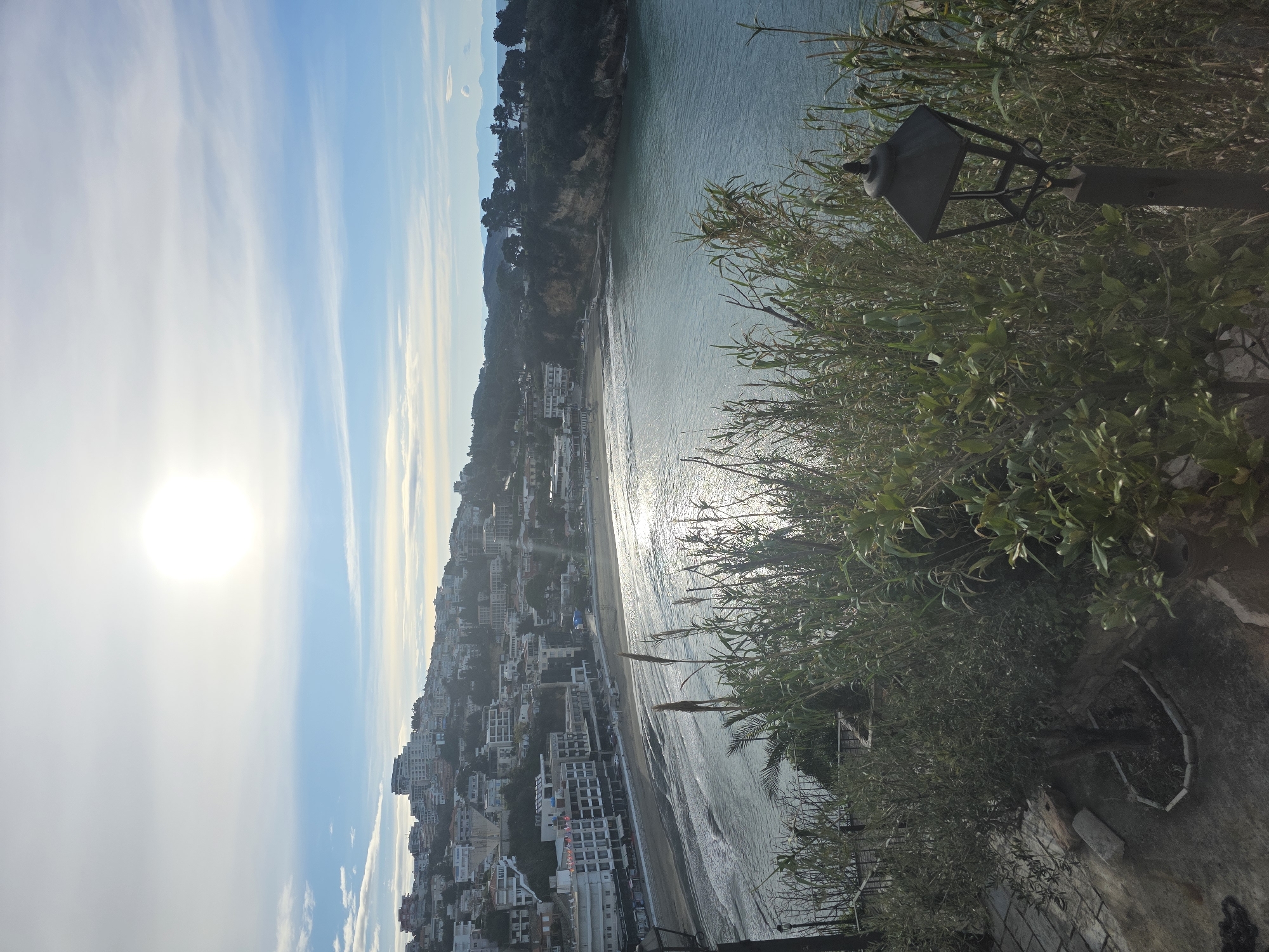 The image depicts a coastal scene in Ulcinj - Ulqin, Montenegro. A beach town is visible, situated along the shoreline, densely populated with buildings. The sea reflects sunlight. Vegetation is prominent in the foreground, along with a lamppost, obscuring a portion of the view. The sky is bright with the sun visible.