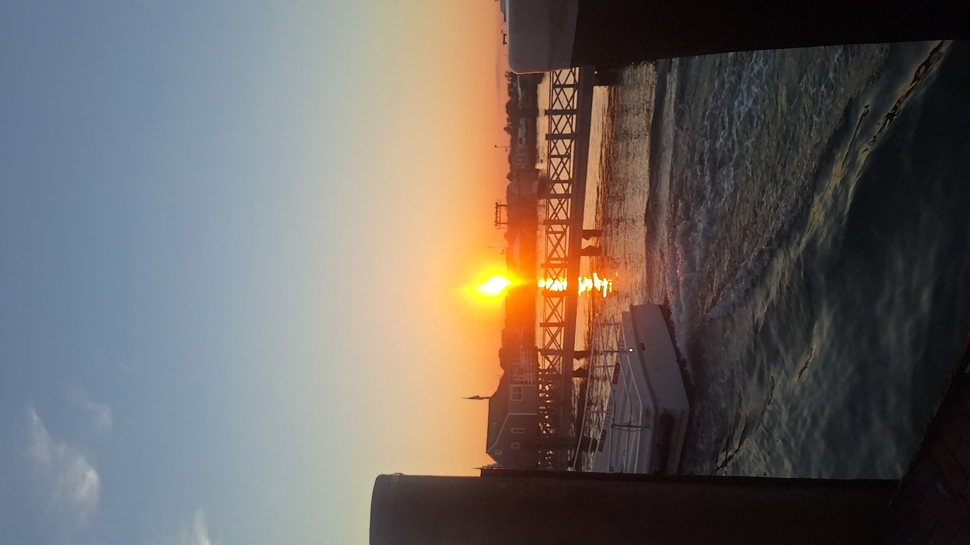 This image captures a sunset over Nantucket Harbor in Nantucket, Massachusetts, USA. The sun, a bright orb with a strong lens flare, is positioned above the horizon, casting a warm orange glow across the sky and water. A wooden pier with crisscrossing railings extends into the harbor, with buildings visible in the background. A white boat with a railing is docked in the foreground, its wake creating ripples on the water's surface. The sky is a gradient of blue to orange, with a few wispy clouds. The scene appears to be a tranquil evening at the harbor.