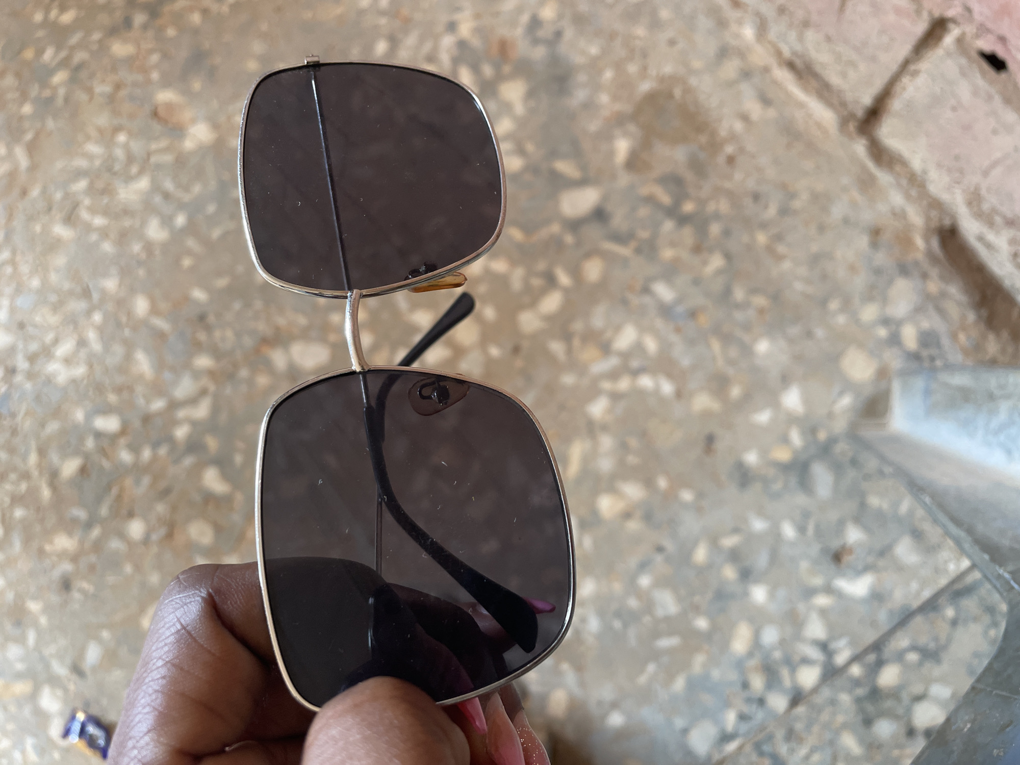The image shows a close-up of a person's hand holding a pair of square-shaped, dark-lensed sunglasses with a thin, gold-toned metal frame. The sunglasses are held in the foreground, with the background consisting of a speckled, terrazzo-like floor or wall, appearing to be outdoors or in an unfinished interior space. The ground is made up of small, irregular-shaped stones and concrete. There are no discernible buildings or specific city landmarks visible to indicate the location as Television, Nigeria. The lighting suggests it is daytime.
