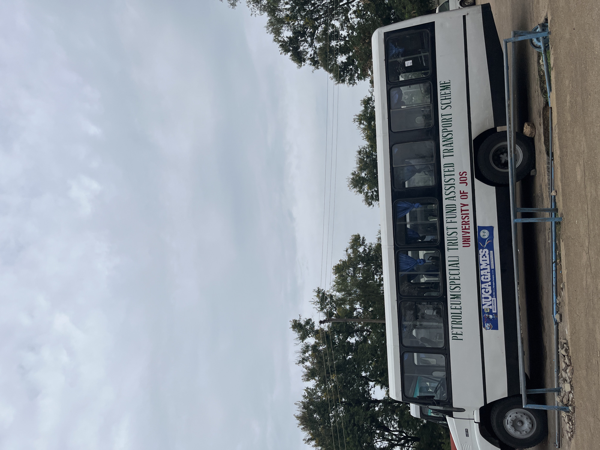 This media file depicts a white bus parked on an asphalt surface, likely in Jos, Nigeria, given the text on the bus. The bus is oriented vertically in the image, with its side facing the viewer. It appears to be a public transport or university bus, as evidenced by the text "PETROLEUM (SPECIAL) TRUST FUND ASSISTED TRANSPORT SCHEME" and "UNIVERSITY OF JOS" printed on its side. There is also a logo with the text "NUGAGAMES" and smaller text underneath, possibly indicating the event or organization associated with the bus.
The bus has several windows, tinted or reflecting the overcast sky, obscuring the interior details. A blue railing or fence is visible along the side of the bus, bordering the parking area. The ground beneath and around the bus is a mix of asphalt and some debris, including small stones and dirt.
In the background, lush green trees with dense foliage are visible, suggesting a potentially natural or campus setting. The sky is cloudy and overcast, indicating a diffused light condition, possibly indicative of an early morning, late afternoon, or a day with inclement weather. The overall mood is neutral, with no visible people or discernible activity taking place. The image is characterized by its focus on the bus and the text displayed on it, serving as a clear identifier of its purpose and affiliation.