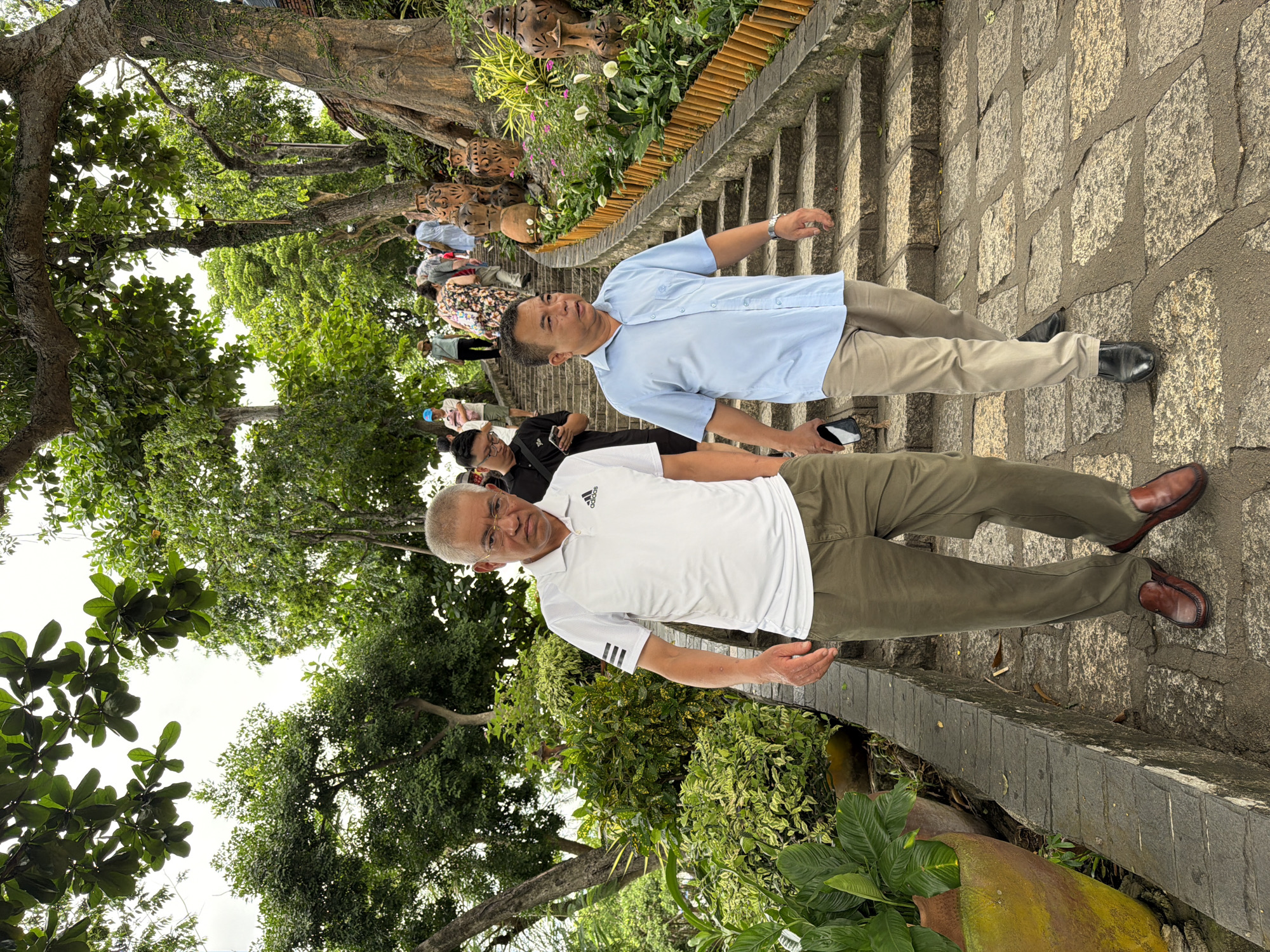 The image depicts a stone staircase leading uphill, lined with lush greenery and large decorative pottery. Several people are ascending the stairs, dressed in casual attire. In the foreground, two men are walking down the stairs. The man on the left wears a white polo shirt with black stripes on the sleeves and olive-green pants. The man on the right is in a light blue short-sleeved shirt and light beige pants. The scene appears to be a tourist site or a temple complex, likely in Nha Trang, Vietnam, given the tropical vegetation and the presence of decorative pottery often seen in Southeast Asian cultural sites. The sky is overcast, suggesting a humid or rainy day.