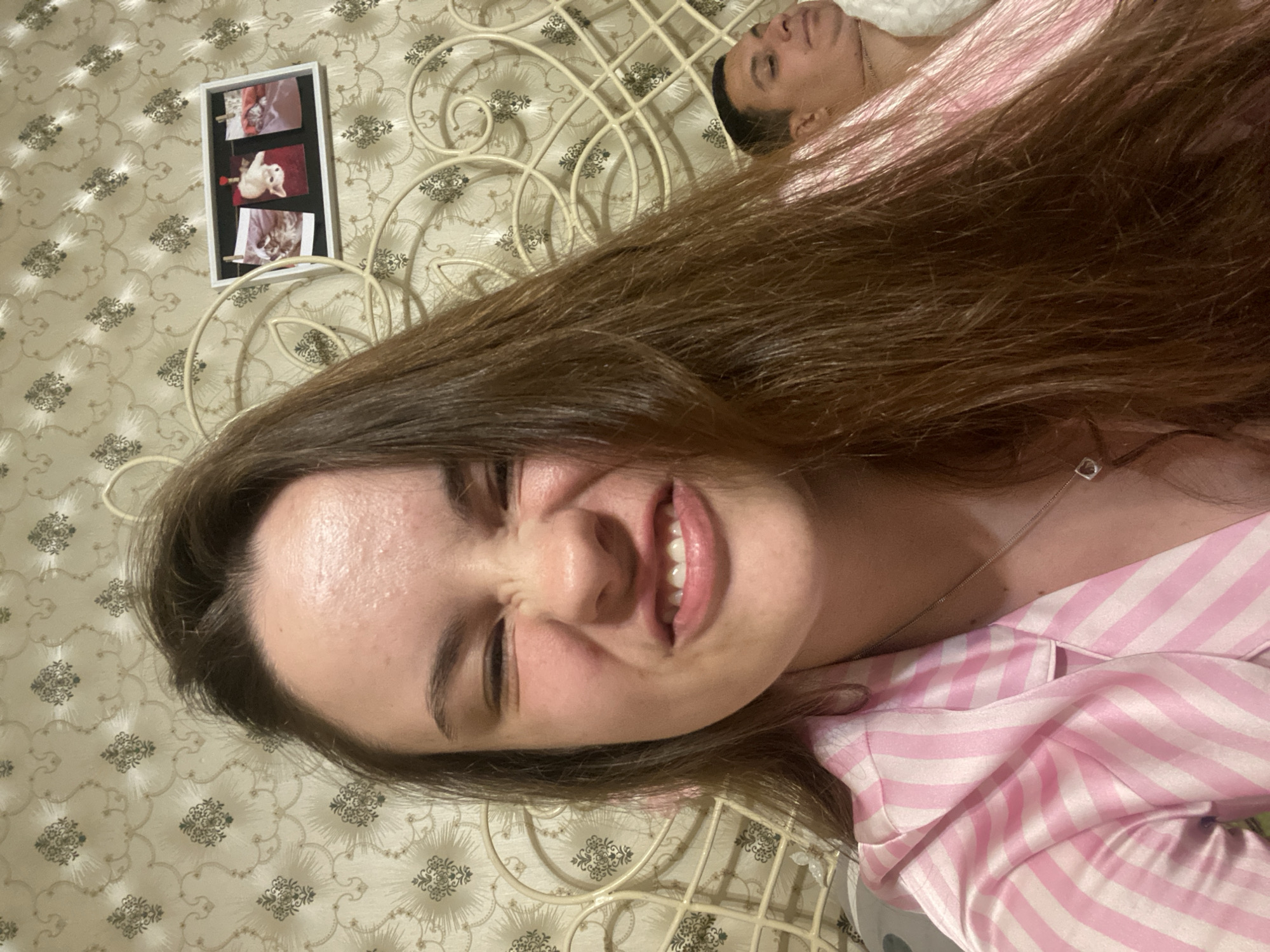 The image is a selfie taken indoors, likely in a bedroom, with a woman in the foreground making a face and a man visible in the background. The woman has long brown hair and is wearing pink and white striped pajamas. Her eyes are squinted, and her teeth are bared in a grimace. She is wearing a silver necklace with a small pendant. The man in the background appears to be lying down and is partially obscured by the woman's hair. He has short dark hair and is wearing a white top.
The background wall is covered with patterned wallpaper. There is a white metal bed frame visible behind the woman. On the wall, to the right of the woman's head, there is a framed picture containing three smaller photos of cats. The lighting suggests an indoor setting at night or with artificial light. The overall scene depicts a casual, personal moment.
The provided information specifies the location as Poltava, Ukraine. However, there are no visual cues in the image that directly identify the city of Poltava, Ukraine. The scene is typical of a domestic interior and lacks any distinct landmarks or indicators specific to that geographical location.