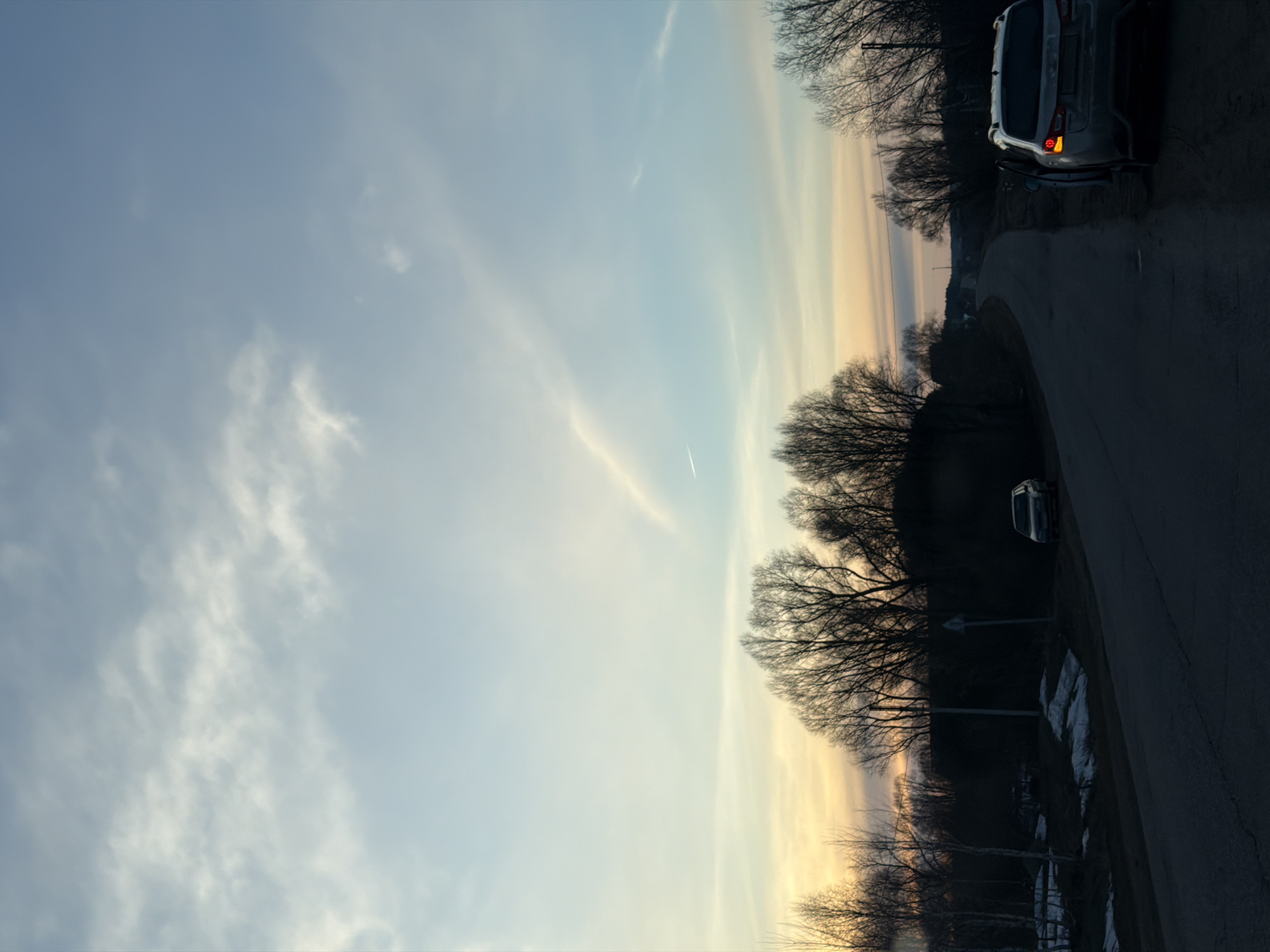 The image shows a partially cloudy sky with a road and trees to the right, a car on the road, and a patch of snow beside it. The setting appears to be at dusk or dawn given the color of the sky. Location could not be identified based on image alone.