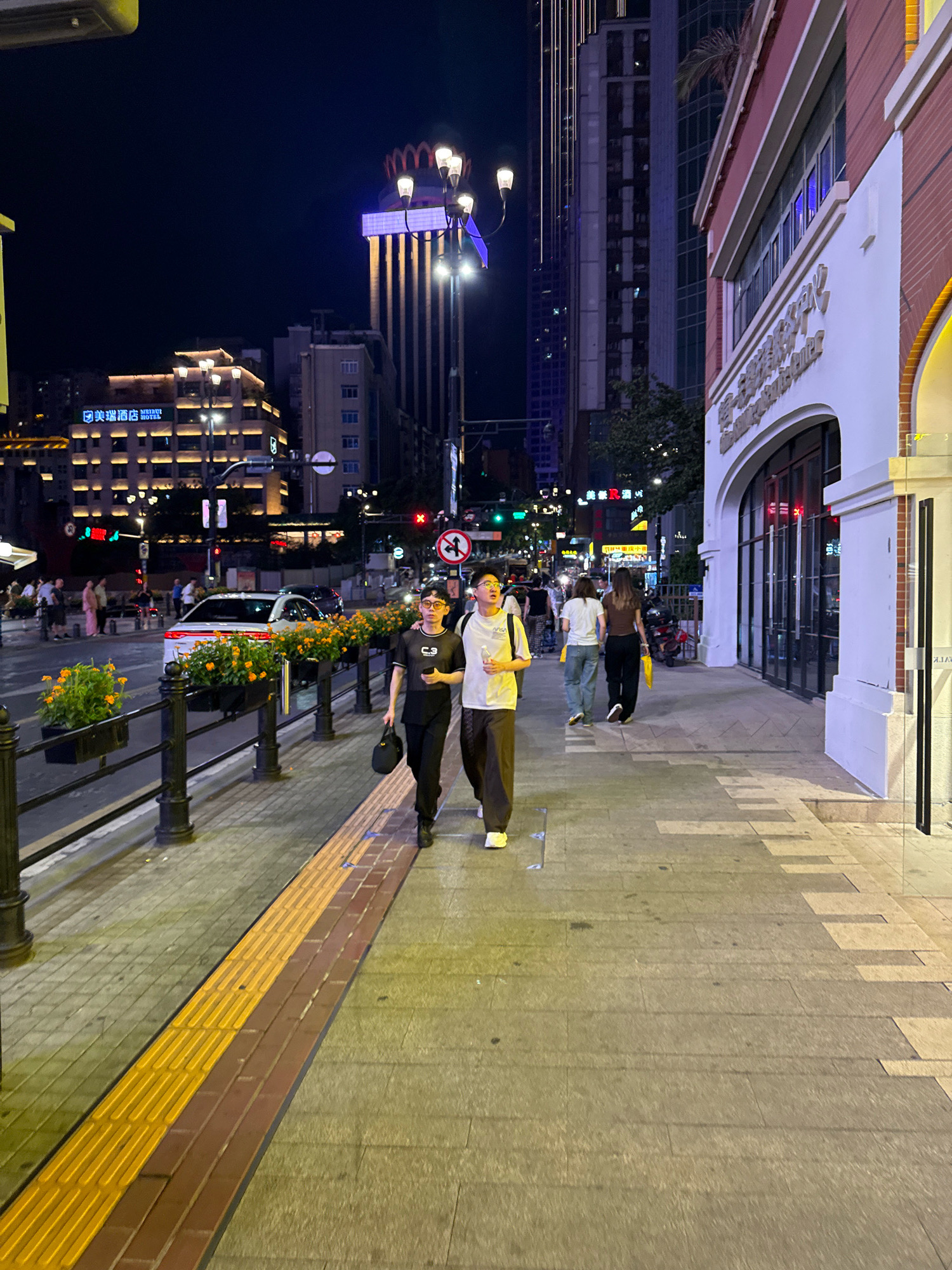 In Yuzhong District, China, at night, two young adults are walking away from the camera on a well-lit sidewalk. The man, in the foreground, wears glasses, a black t-shirt with a white graphic, and dark pants, carrying a black bag. The woman beside him wears a white t-shirt and dark pants. They are walking past a row of black planters filled with orange flowers, separated from the street by a black metal railing. Beyond the railing, a white sedan is parked, and further in the background, the city skyline is illuminated by numerous lights, with tall buildings and commercial signage visible, including "MEIRUI HOTEL" in English. The sidewalk is paved with textured tiles, including a yellow tactile paving strip for the visually impaired. The overall scene depicts a typical urban environment at night.