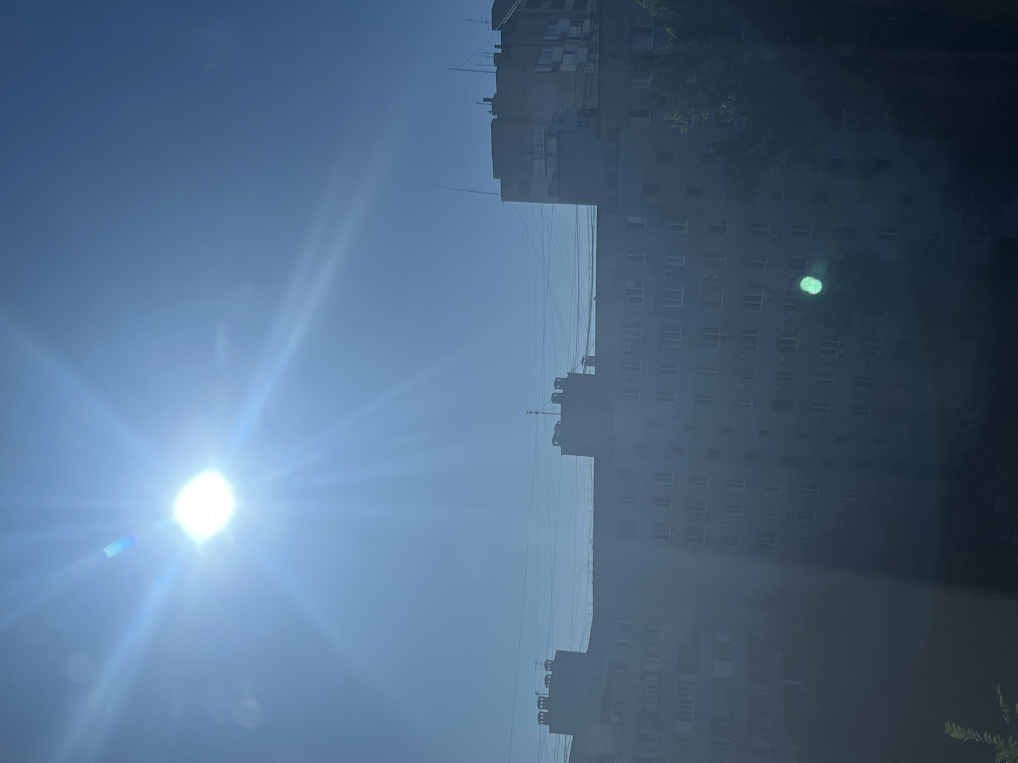 The image depicts a bright, sunny day in Volgograd, Russia, with the sun directly overhead, casting lens flare across the frame. In the foreground, the silhouette of a large, grey apartment building is visible, characterized by numerous windows and what appear to be air conditioning units. The building's architecture suggests it is a typical Soviet-era residential block. Overhead, a tangle of power lines stretches across the clear blue sky. Another, taller apartment building is partially visible on the right side of the frame. The overall impression is of a typical urban landscape on a clear day, with the intense sunlight creating a slightly hazy or washed-out effect on the buildings. There are no people or vehicles visible in the image.
