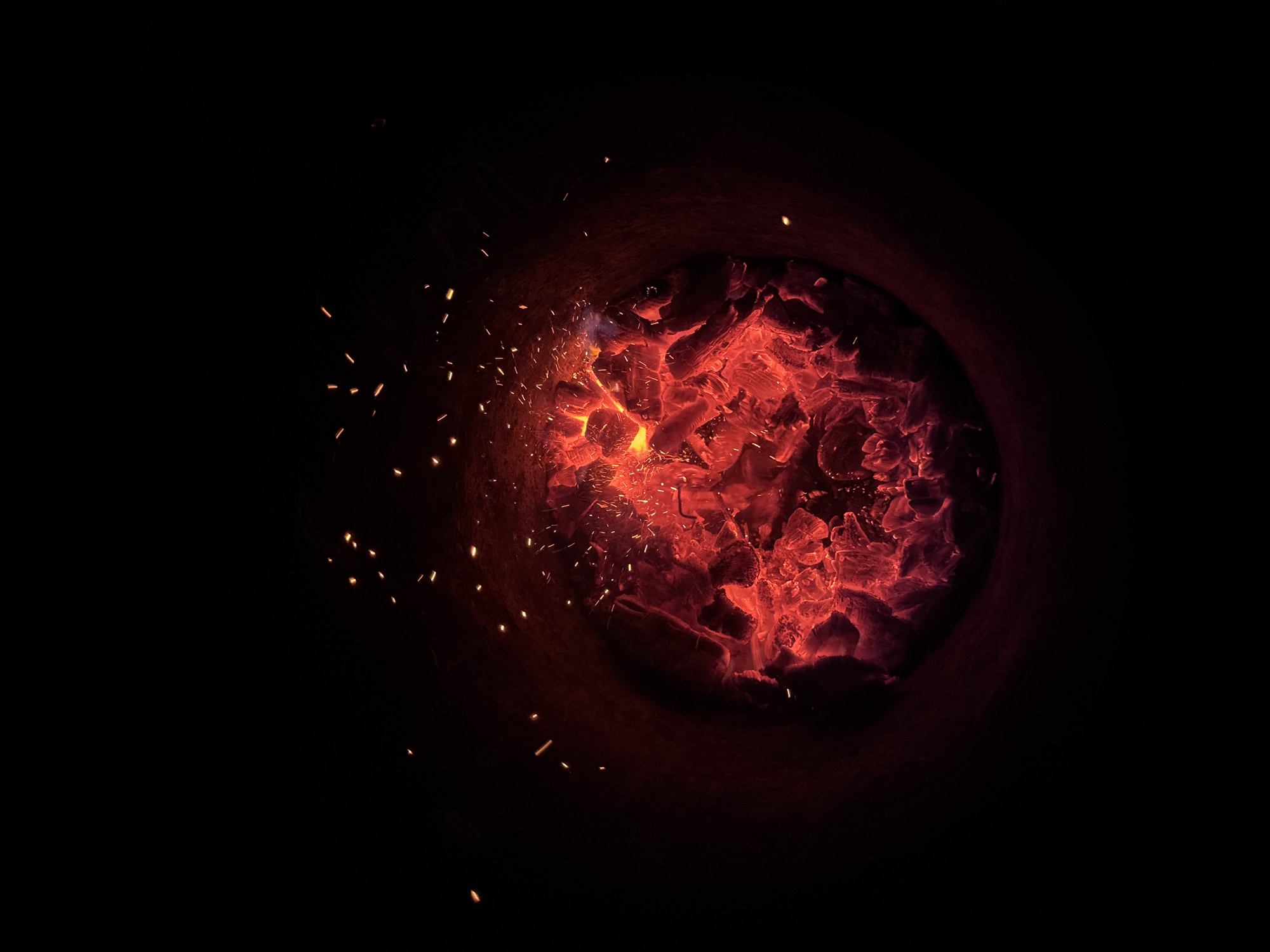 This image depicts a close-up view of glowing red embers and sparks from a fire, viewed from above within a dark circular opening, likely a fire pit or brazier. The coals are intensely hot, emitting a vibrant red and orange light that illuminates the surrounding darkness. Small, bright sparks are scattered in the air above the embers, indicating an active fire. The scene is abstract and focuses on the intense heat and light of the burning material. There is no discernible location in the city of Chaikovsky, Russia, or any people or specific objects identifiable beyond the embers themselves. The environment is characterized by deep shadows and the glowing focal point of the fire.