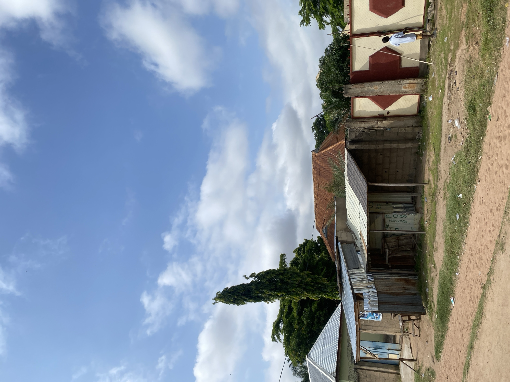 The image depicts a street scene in Jauro Yinu, Nigeria, under a bright blue sky with scattered white clouds. In the foreground, a dirt path leads to a row of simple buildings. The structures are constructed with a mix of materials, including corrugated metal roofing, cinder blocks, and what appears to be mud or plaster walls. Some of the buildings have open fronts, revealing tables and shelves, suggesting they might be small shops or market stalls. One building has a sign that reads "POS HERE".
To the right of the buildings is a large gate, painted cream and burgundy with a geometric diamond pattern. A young man in a light blue shirt and yellow shorts is standing near the gate, holding a long, light-colored stick, seemingly interacting with the gate or the wall. Behind the buildings and gate, lush green trees and foliage are visible. A tall, slender evergreen tree stands out prominently among the other trees. The overall impression is of a modest, working-class area on a sunny day.