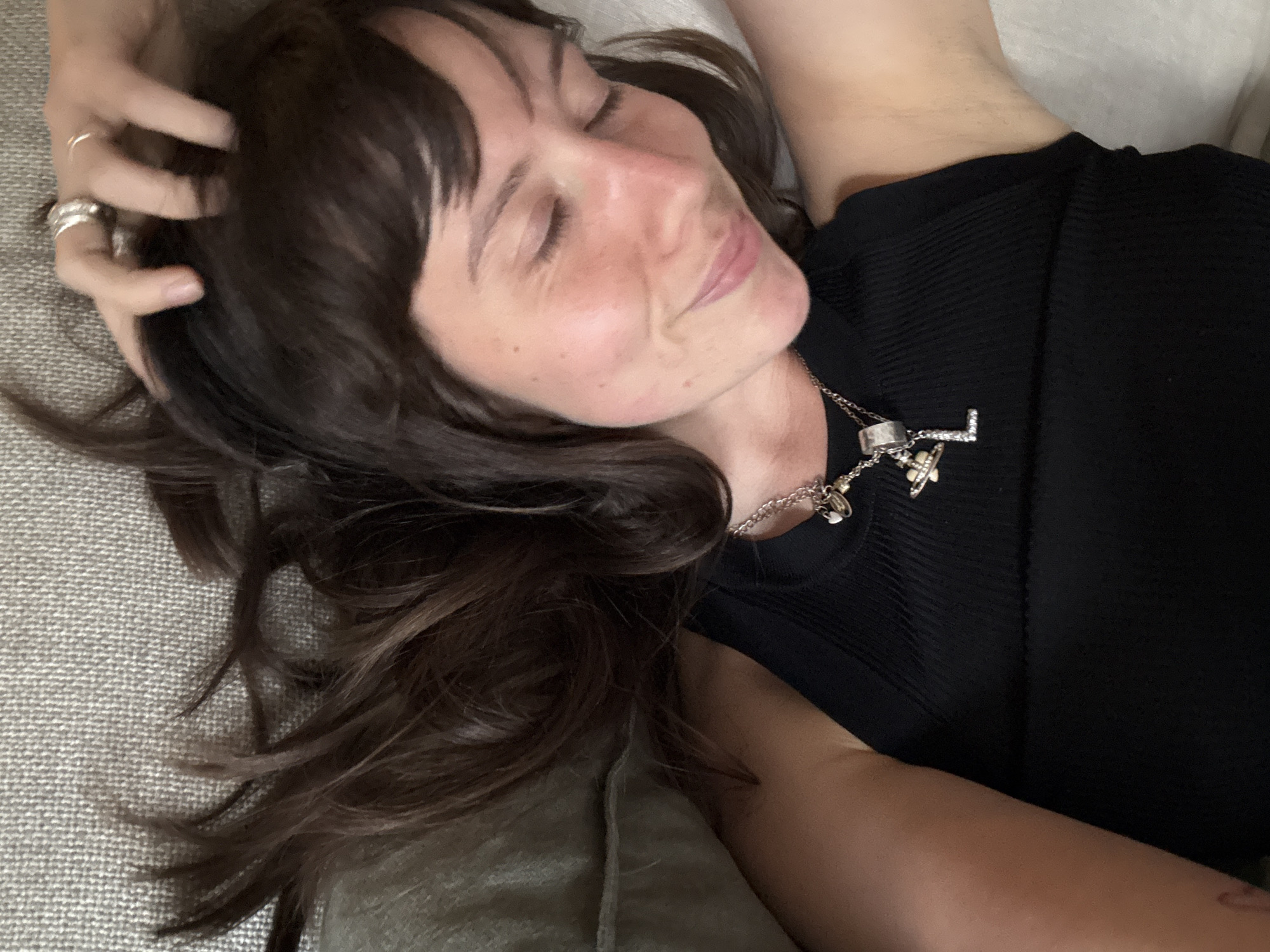 A medium close-up shot captures a person, likely female, resting comfortably in what appears to be an indoor setting. The individual is shown from the chest up, lying on a light grey, textured fabric surface, possibly a sofa or bed. Their head is tilted slightly, and their eyes are closed, with a soft, contented smile playing on their lips, suggesting a state of relaxation or peacefulness.
The person has dark brown hair with bangs, which falls around their face and spreads across the textured surface. One hand, adorned with several silver rings on the fingers, rests near their head, gently touching their hair. They are wearing a black ribbed top or dress. A silver chain necklace is visible around their neck, featuring multiple charms, including a distinctive silver letter 'L' embellished with small stones, and another charm resembling a stylized star or cross.
The lighting is soft and even, indicating an indoor environment, possibly during the day or early evening. The background is muted, with hints of a lighter fabric behind the head and an olive green/darker fabric in the lower left, contributing to a calm and intimate atmosphere. No explicit text is visible besides the letter 'L' on the necklace charm. The overall impression is one of quiet repose, likely within a home in London, United Kingdom.