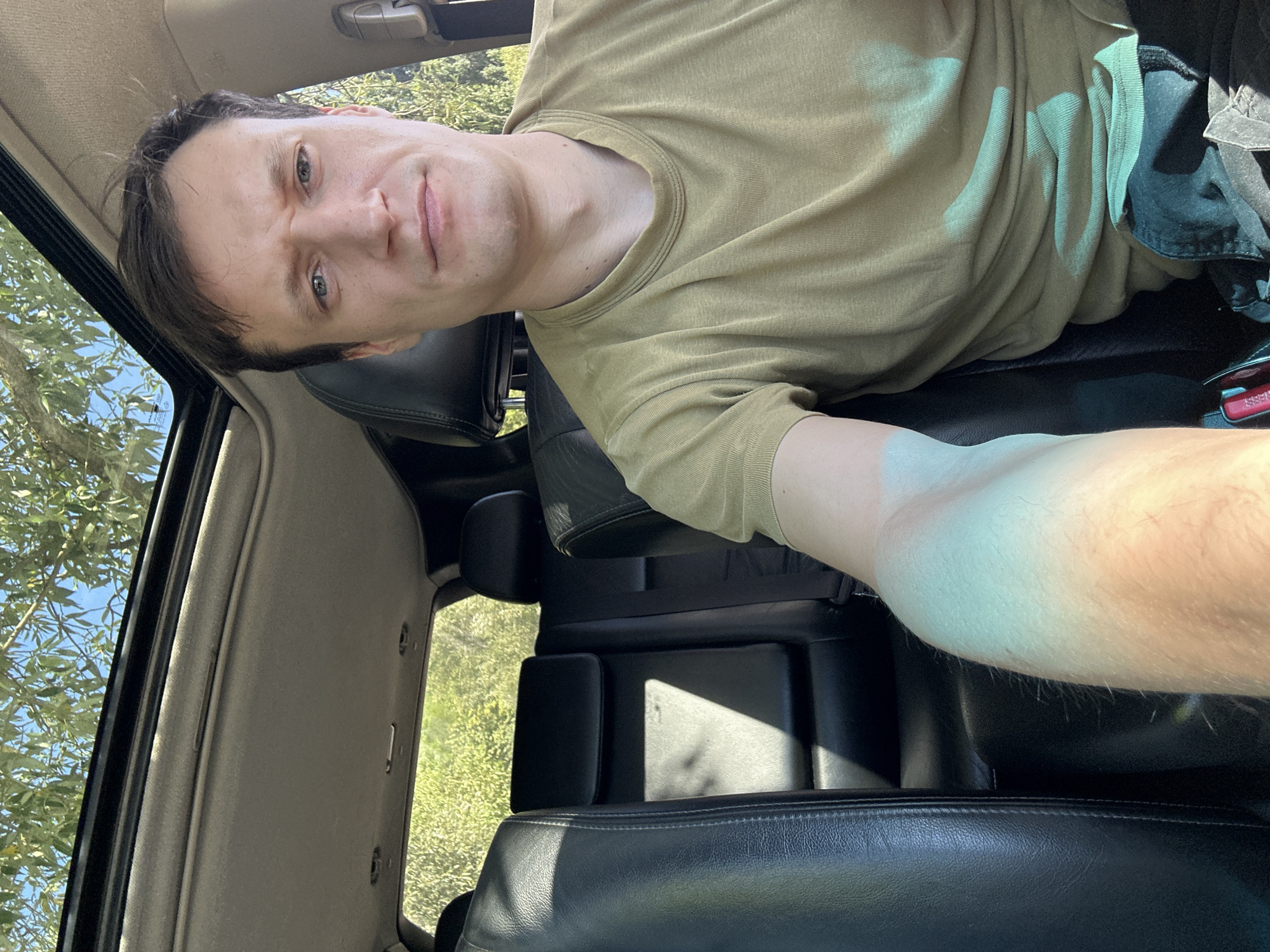 A young man is seated in the front passenger seat of a dark-colored SUV, facing the camera. He has short, dark brown hair and fair skin. He is wearing a light olive-green t-shirt. Sunlight streams through the open sunroof of the vehicle, casting shadows on the black leather interior and the man's arm. The background visible through the sunroof and rear window consists of green foliage and blue sky, suggesting an outdoor setting, possibly near Makiv, Ukraine. The man's expression is neutral, and his left arm is extended forward, partially obscuring the view of the backseat.