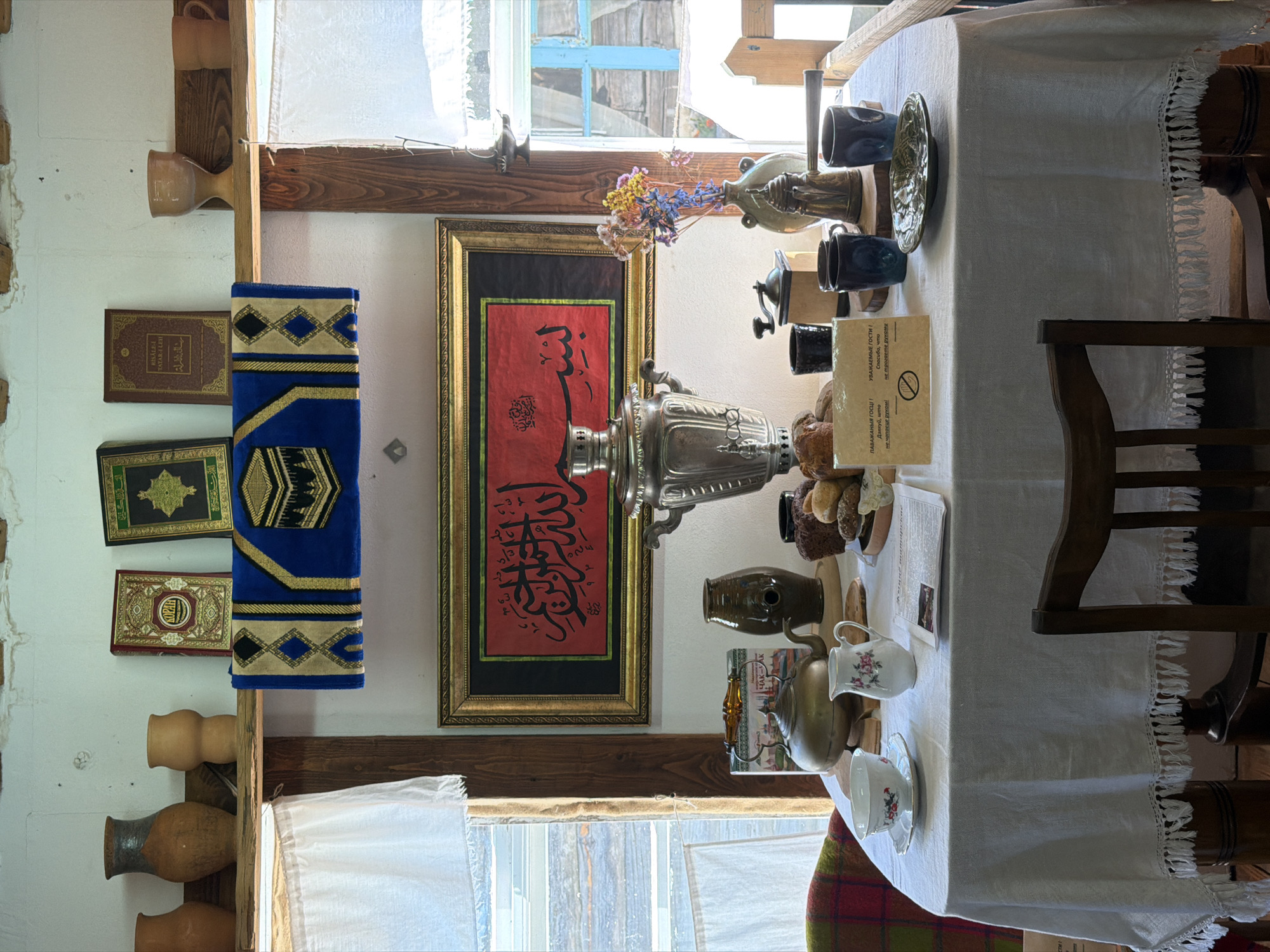 The image depicts a still life scene on a table set inside what appears to be a rustic room. The table is covered with a white linen tablecloth adorned with fringe. On the table, various objects are arranged, suggesting a tea or coffee service and some food items. These include a silver samovar, a silver teapot, a ceramic pitcher with floral patterns, a teacup and saucer with floral designs, and a small stack of bread or pastries. There are also several dark blue mugs and a small decorative plate. A newspaper or leaflet is laid out near the pitcher, and a sign with text in Cyrillic script is positioned on the right side of the table.
Above the table, a framed piece of artwork with red background and black calligraphic script is hung on the wall. To the left of the artwork, a blue rug with gold and black geometric patterns is displayed, featuring an embroidered image of the Kaaba. On a shelf above this rug, several objects are visible: three books, likely religious texts given their appearance, and several clay vases and vessels of varying sizes and shapes. One book is red with gold lettering, another is dark with gold ornamentation, and a third appears to be a brownish-red volume.
The room itself seems to have wooden beams and a white-painted wall, with natural light streaming in from windows on the left and right, which have white curtains. The window on the right shows a glimpse of blue wooden structures and vegetation outside, suggesting a rural or semi-rural setting.
Based on the provided information, this scene is located in Litvenski Rural Council, Belarus. The overall impression is one of a cultural display, possibly within a museum, historical house, or traditional home, showcasing elements of local heritage, and potentially Islamic traditions given the presence of the Quranic calligraphy, Kaaba imagery, and books. There are no people visible in the image.