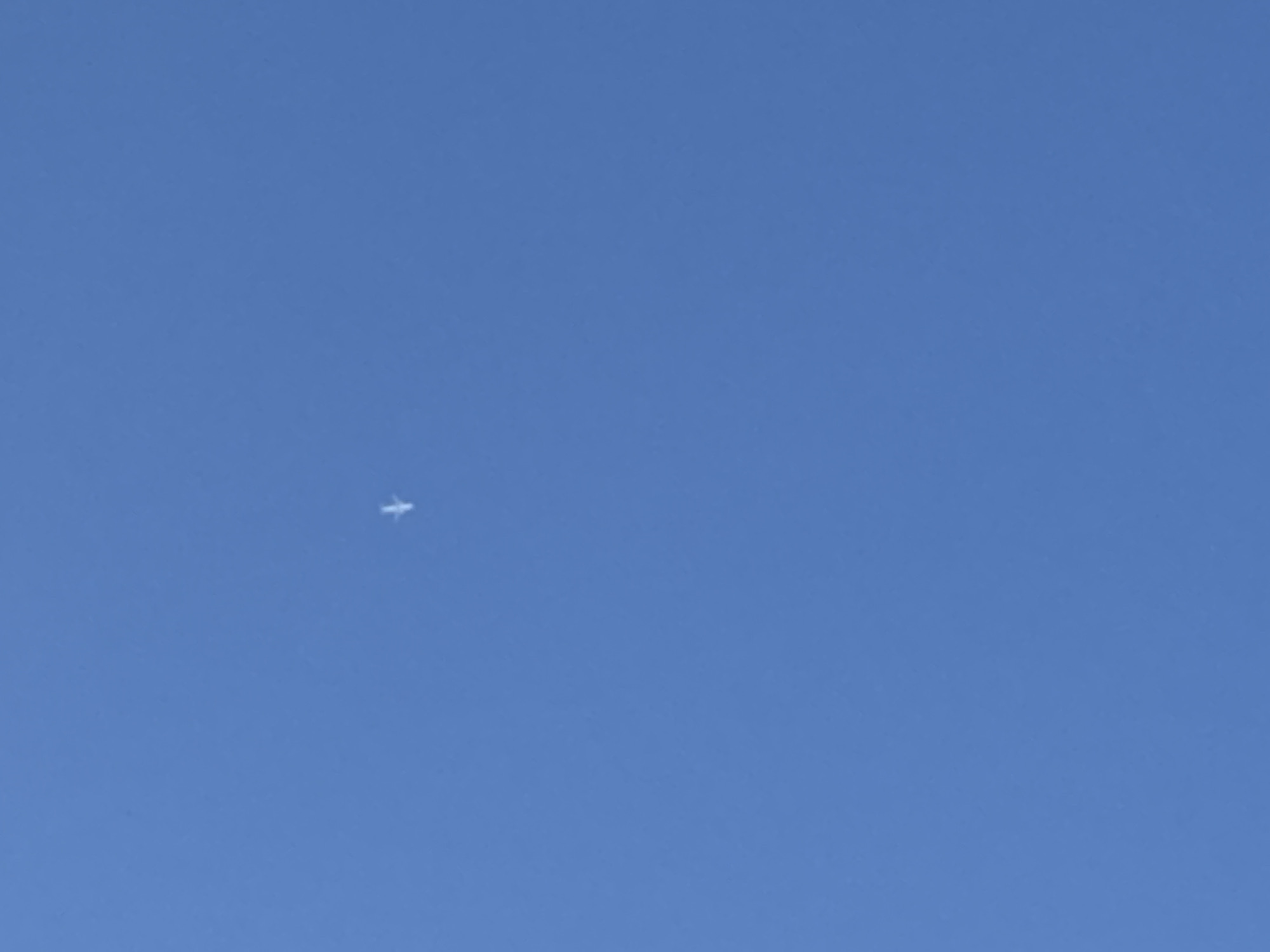A single airplane is visible in the center of a clear blue sky. The airplane is small and appears to be white. The image is a photograph of the sky with no other objects or people present. The location is described as Novi Kiner, Russia, though this information cannot be verified from the image itself.
