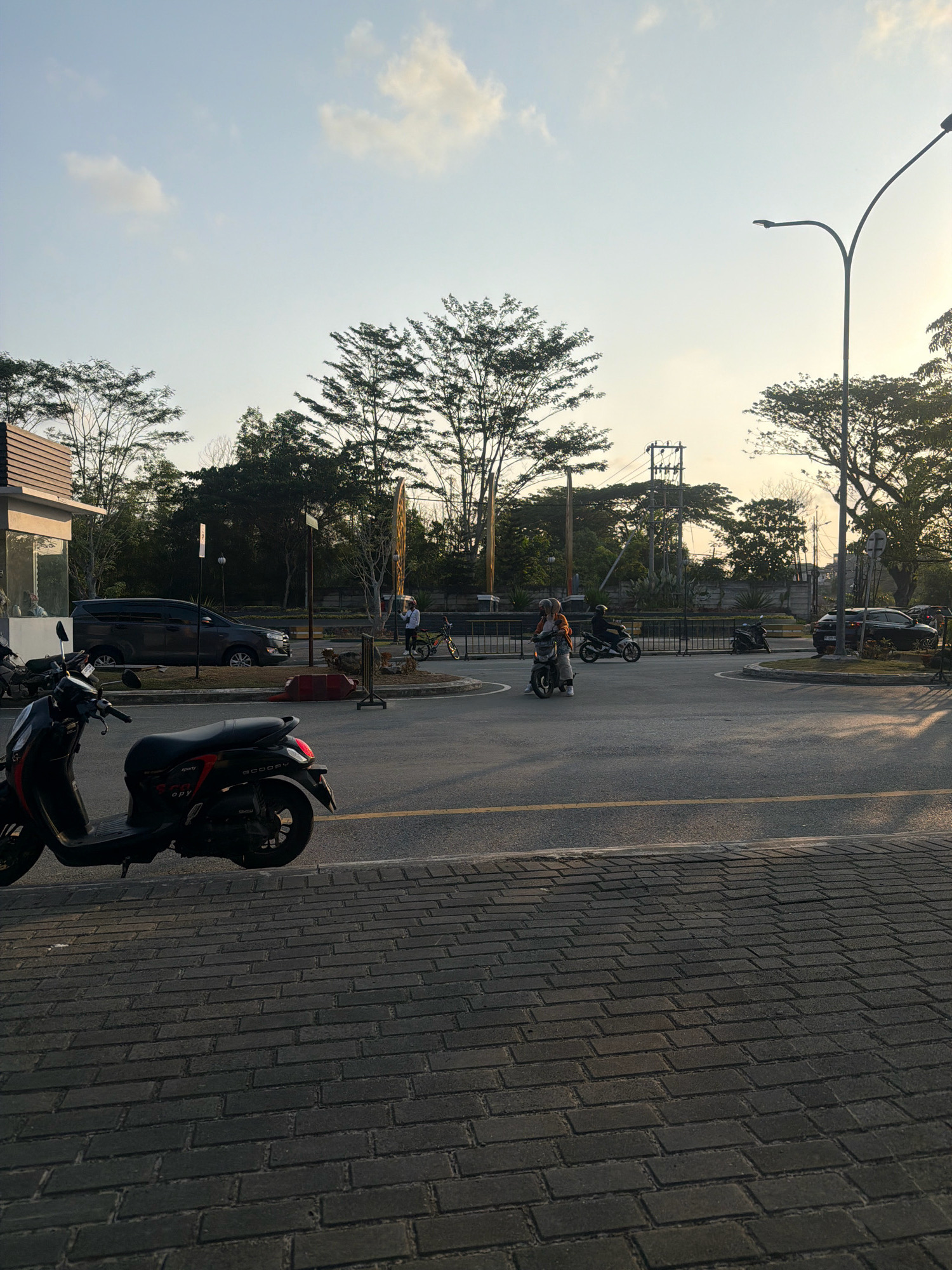 The image depicts a street scene in Balikpapan, Indonesia, during what appears to be late afternoon or early evening, judging by the warm lighting and long shadows. In the foreground, a dark-colored scooter is parked on a brick-paved area adjacent to a paved road. The road has a yellow line marking the lane division.
Further into the scene, a road intersection is visible. Several people on motorcycles are navigating the intersection. One person on a scooter, wearing a light-colored helmet and an orange top, is moving towards the left. Another person on a dark motorcycle is behind them. A dark-colored SUV is parked to the right of the intersection.
On the left side of the image, a modern building with large glass windows and a brown, slat-like facade is partially visible. Trees line the background, some with sparse foliage, suggesting a tropical climate. A street lamp with a modern design arches over the road on the right side of the image. Electrical poles and wires are also visible in the mid-ground, indicating the presence of infrastructure. The sky is mostly clear with a few scattered clouds, illuminated by the soft glow of the setting sun. The overall impression is a typical urban street in a tropical city.
