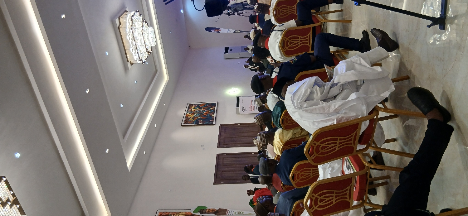 In a brightly lit hall located in Tudun Nupawa, Nigeria, an audience is seated in red and gold chairs, attending what appears to be a formal event. Many of the attendees are dressed in traditional attire. The hall features a chandelier, artwork on the walls, and visible doors. A microphone stand is present.